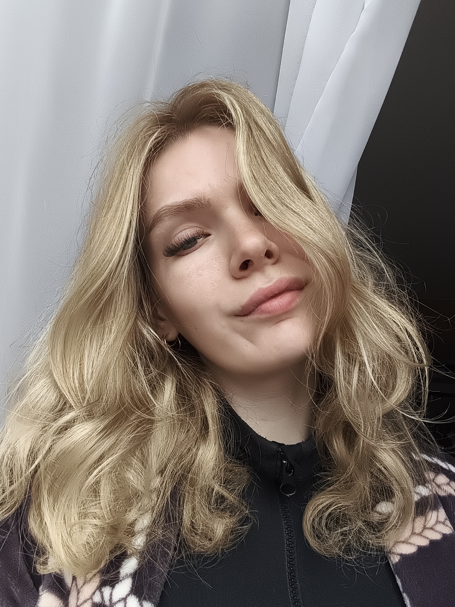 The image shows a selfie of a fair-skinned young woman with long, wavy blonde hair partially covering her face. She is wearing a dark-colored zippered jacket. Behind her is a white curtain partially obscuring a dark area. The location in the city of Русский Вожой, Russia is unknown and cannot be determined from the image.