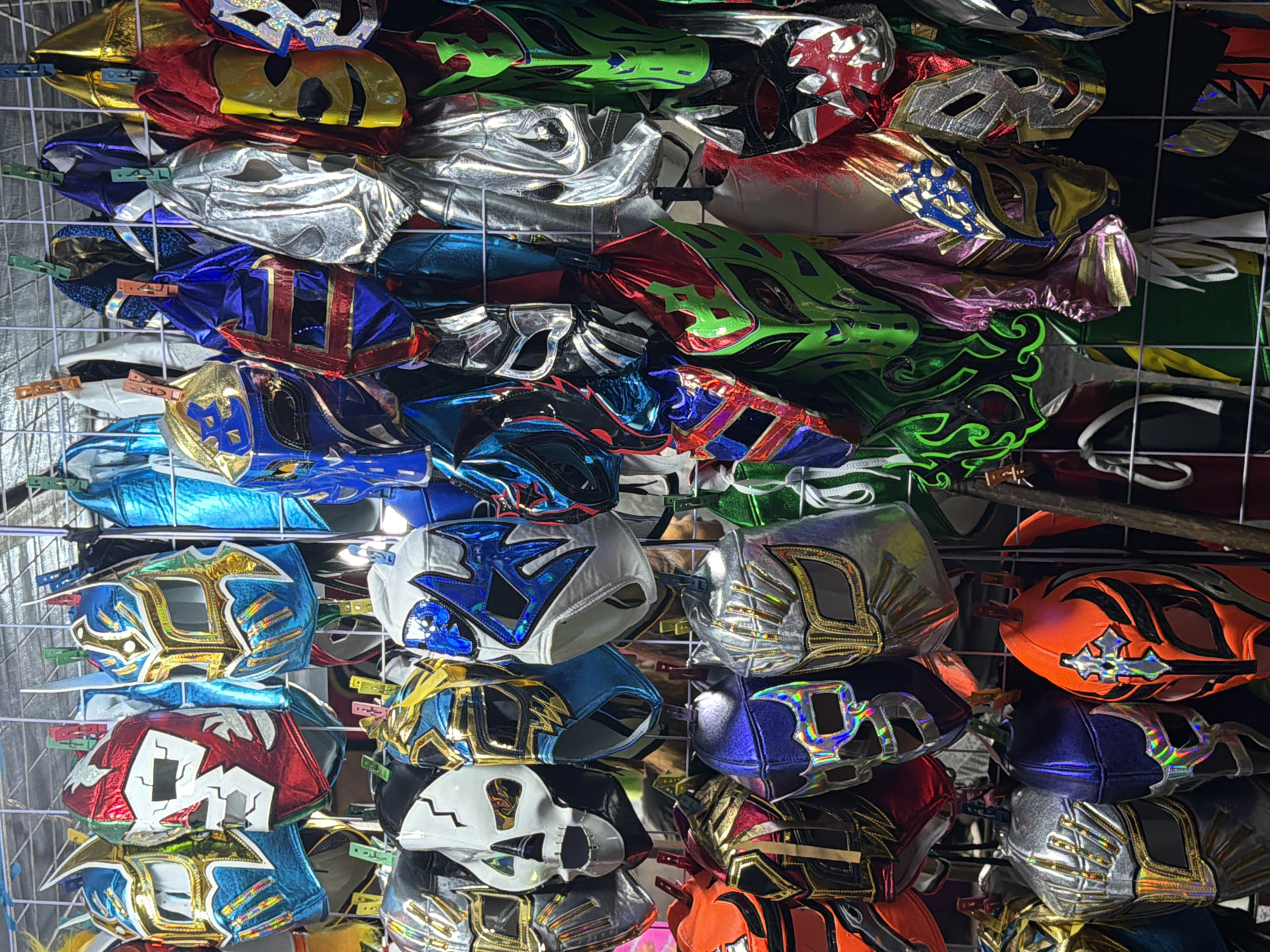 A collection of Lucha Libre wrestling masks is displayed on a metal wire rack. The masks are arranged in rows and columns, each attached to the rack with clothespins. The masks vary in color and design, with many featuring metallic accents and intricate patterns. The location is Puebla, Mexico.