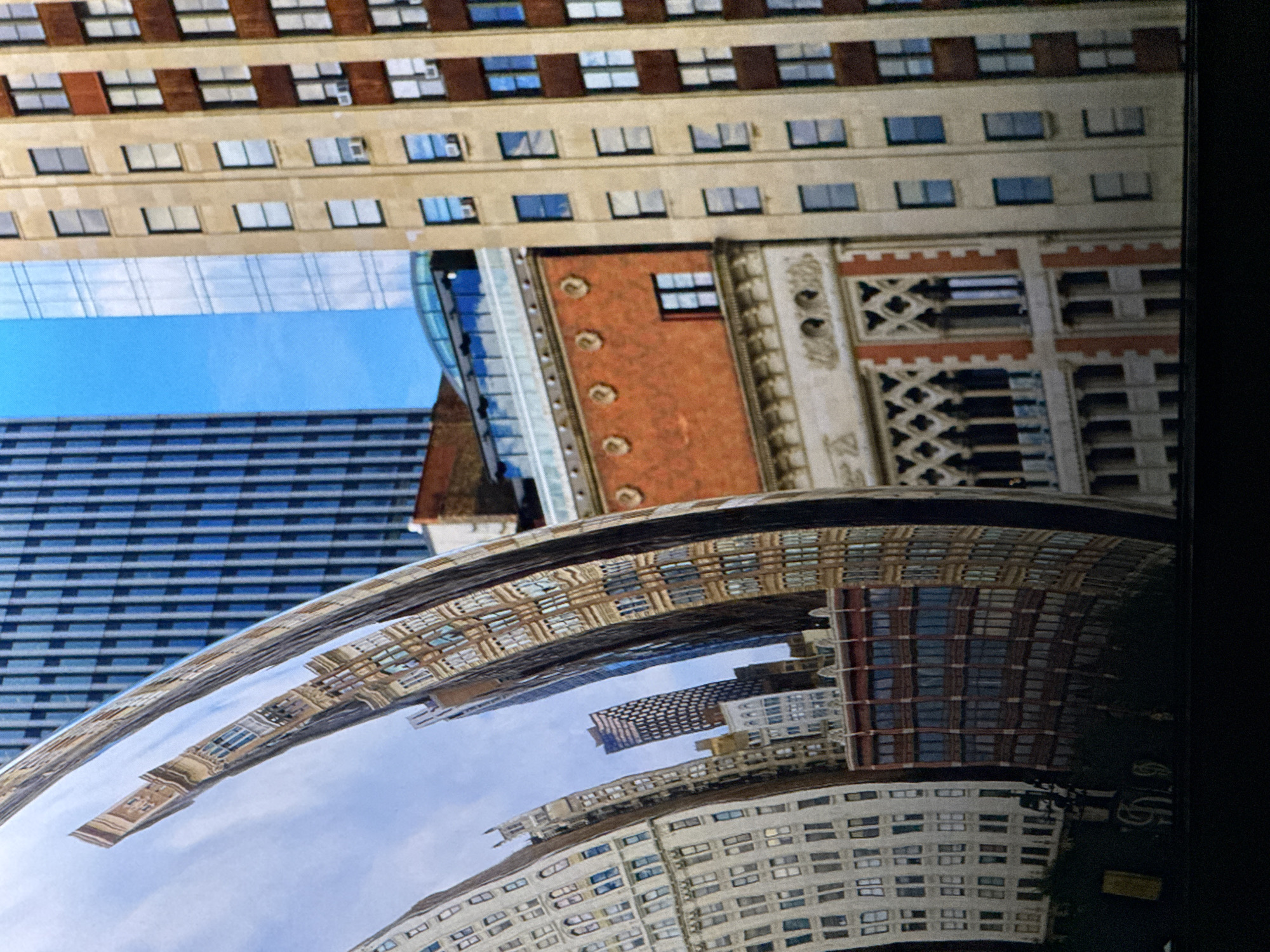 This is a close-up, high-angle, exterior shot of the reflections on the Cloud Gate sculpture in Chicago, Illinois. The sculpture, made of polished stainless steel, distorts the reflections of the surrounding skyscrapers. The buildings are a mix of modern and older architecture, with glass and brick facades. The sky is visible, a bright blue with some clouds. The reflections are warped and stretched, giving the impression of a fluid, dynamic cityscape. The foreground of the image is dark, suggesting the sculpture is at street level.