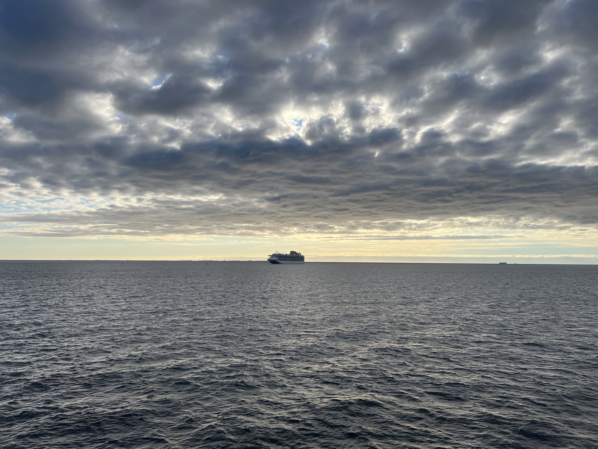 The image shows a wide view of the sea under a cloudy sky. In the center of the frame, a large white cruise ship is visible on the horizon. The sea is a dark blue, with visible waves. The sky is filled with gray and white clouds, with patches of sunlight breaking through. The horizon line is flat and even. There is a small buoys in the water. In the far distance, faint landmasses or other ships are barely visible. The overall atmosphere is calm and serene. The location is the sea off the coast of Gdynia, Poland.