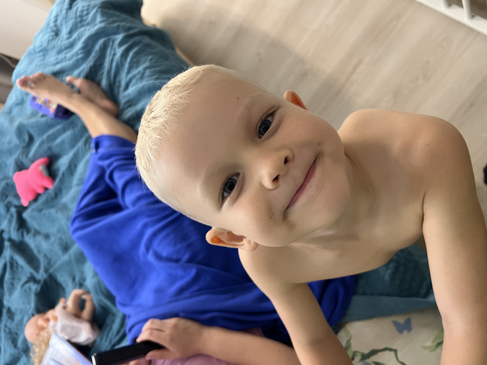 An overhead, close-up shot captures a candid indoor moment in Sochi, Russia, featuring a young, fair-skinned child with short blonde hair and bright blue eyes smiling directly at the camera. The child, likely a boy, is shirtless, prominently positioned in the center-right of the frame, conveying a cheerful and relaxed emotion.
Below and to the left of the main subject, another person is partially visible, lying on a textured blue blanket. This person's bare feet, clad in pink nail polish, are seen resting on the blanket near a small purple toy. They are wearing a royal blue long-sleeved top and are holding a black object, possibly a smartphone or remote, along with a book. A blonde-haired doll is also visible lying near their hands. A pink plastic toy rests on the blue blanket.
The background shows light-colored wooden flooring in the upper right, and a white object, possibly part of a radiator or furniture, in the top right corner. A patterned pillowcase or sheet with a blue butterfly design is subtly visible under the main child's right arm. The bright lighting suggests it is daytime. The scene evokes a casual and comfortable atmosphere, likely depicting playtime or relaxation at home.