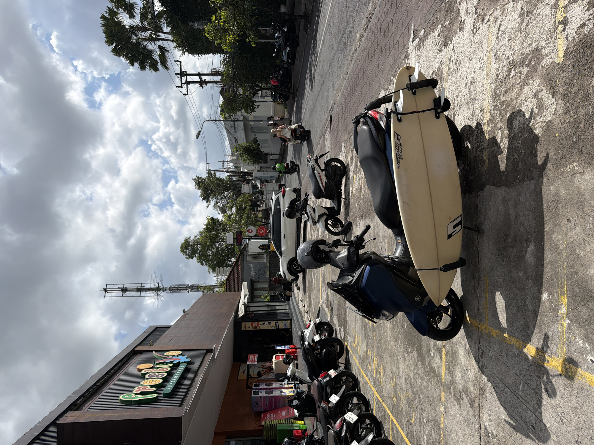 The image depicts a street scene in North Kuta, Indonesia. A row of parked motorbikes lines the edge of a building identified by a sign that reads "Pepito Supermarket". One blue motorbike has a surfboard secured to its side. In the background, other motorbikes are visible on the street. People, likely pedestrians or drivers, are present in the background on motorbikes. The sky is partly cloudy.