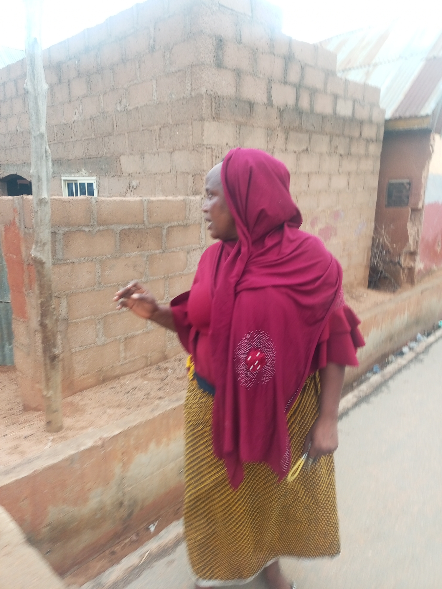 An individual wearing a maroon headscarf and a brown and yellow patterned skirt stands on the side of a street. She is in Tempure, Nigeria, and is positioned next to a beige brick building and a concrete curb. The individual appears to be gesturing with her right hand and holding a yellow object in her left.