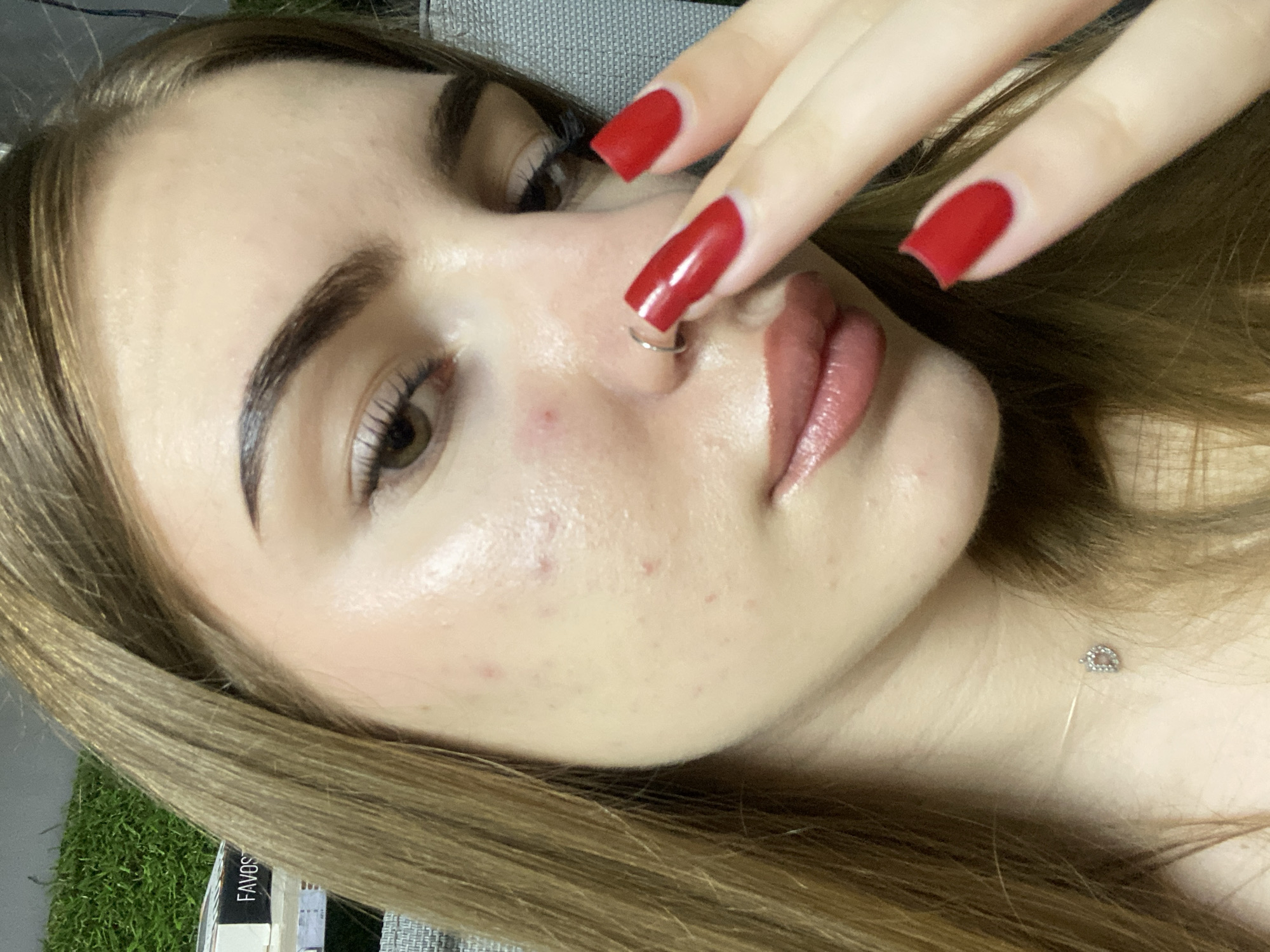 The image shows a close-up of a young woman's face, with a focus on her skin, makeup, and fingernails. Her skin appears to have some blemishes, and she is wearing makeup, including foundation, blush, and lip gloss. Her eyebrows are well-defined, and she has long, dark eyelashes. Her fingernails are painted a bright red, and she has a nose ring. She is wearing a thin silver necklace with a small charm. Her hair is long and brown. The background is blurred, but it appears to be an indoor setting, possibly a room with some greenery or a decorative wall covering. There is a product box with the word "FAVOS" visible on the left side of the image. There are no explicit indications of the location being Дьяково, Russia.