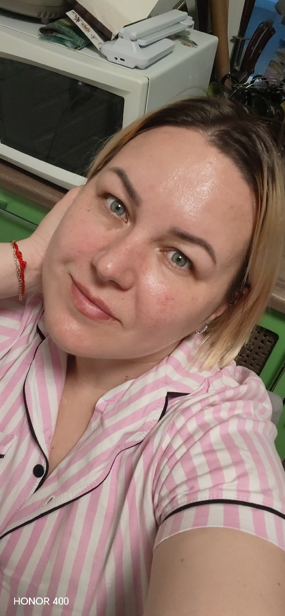 A medium close-up selfie depicts a woman wearing a pink and white striped pajama top with black trim and black buttons. She has fair skin, short blonde hair with darker roots, and blue eyes. The woman is looking directly at the camera with a slight smile. She has a red bracelet on her left wrist. Her right hand is near her face, touching her neck.
Behind her is a white microwave, with what appears to be a white router-like device sitting on top of it. In the background, there are also hints of green and brown objects. The bottom of the image contains the text "HONOR 400". There is no information about the city of Noviy, Russia in the image.