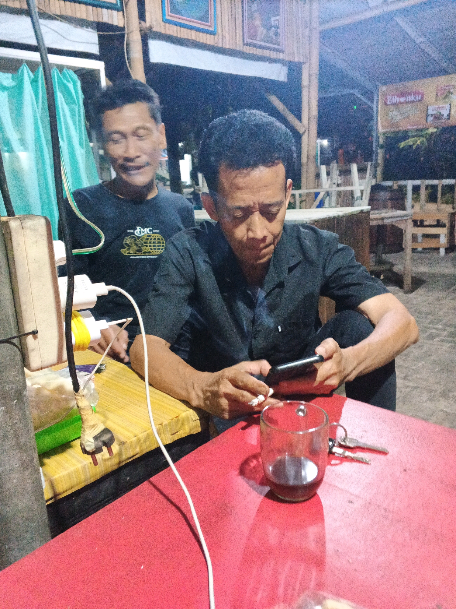 The image captures two men in an outdoor setting, likely a cafe or warung, in Ungaran, Indonesia. The man in the foreground, with short, dark hair, is seated and focused on his smartphone. He is wearing a dark, short-sleeved collared shirt and dark pants. In his left hand, he holds a cigarette. On the red table in front of him, a clear glass mug containing a dark liquid, possibly coffee, and a set of keys are visible. The man in the background, also with short, dark hair, is standing or leaning behind the man in the foreground. He is wearing a dark t-shirt with a graphic on it. The environment appears to be dimly lit, suggesting it is evening or night. Wooden structures and bamboo elements are visible in the background, along with a sign that reads "Bihonku." Electrical outlets and charging cables are present on a table to the left of the frame, indicating a place where people can charge their devices. The ground is paved with bricks or stones. The overall impression is a casual, relaxed scene.