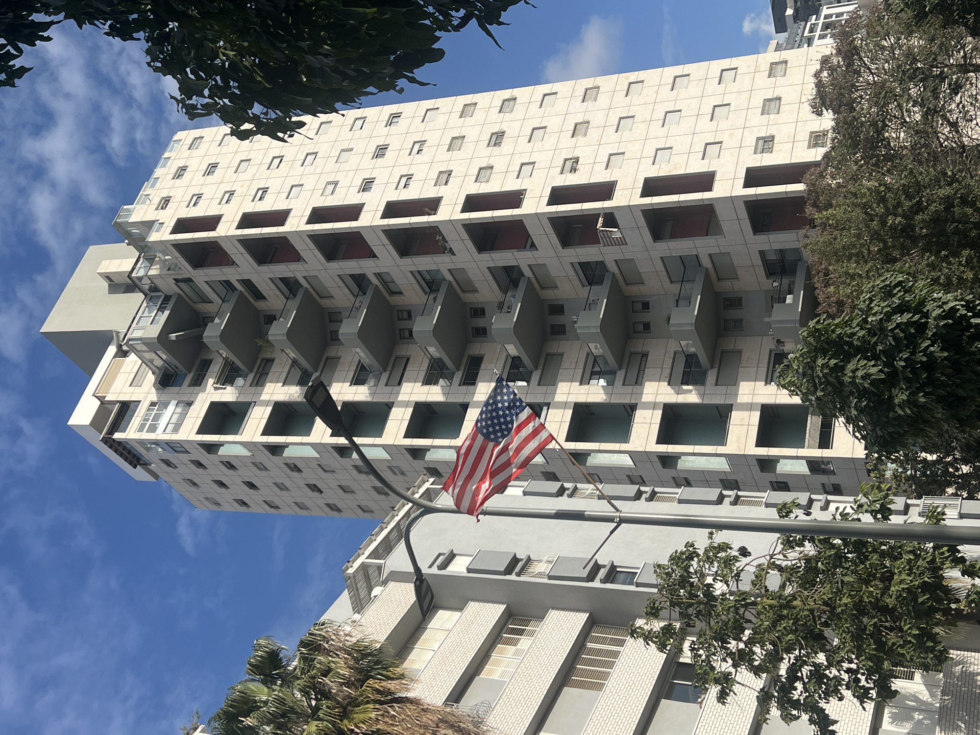 A low-angle daytime shot depicts a multi-story building with a modular architectural design, possibly in Ramat Gan, Israel, although the location is not explicitly confirmed. The building is predominantly white or light beige, featuring recessed balconies or open spaces on several floors. Windows are rectangular and evenly spaced. An American flag is prominently displayed, likely on a flagpole attached to a lower building or structure in the foreground. The sky is partly cloudy, with blue visible through the gaps in the trees framing the top left and right sides of the image. Another building, appearing smaller and in a similar architectural style, is partially visible in the foreground. A street light is visible near the flag. The presence of trees suggests an urban environment with landscaping. No people are visible in the frame.