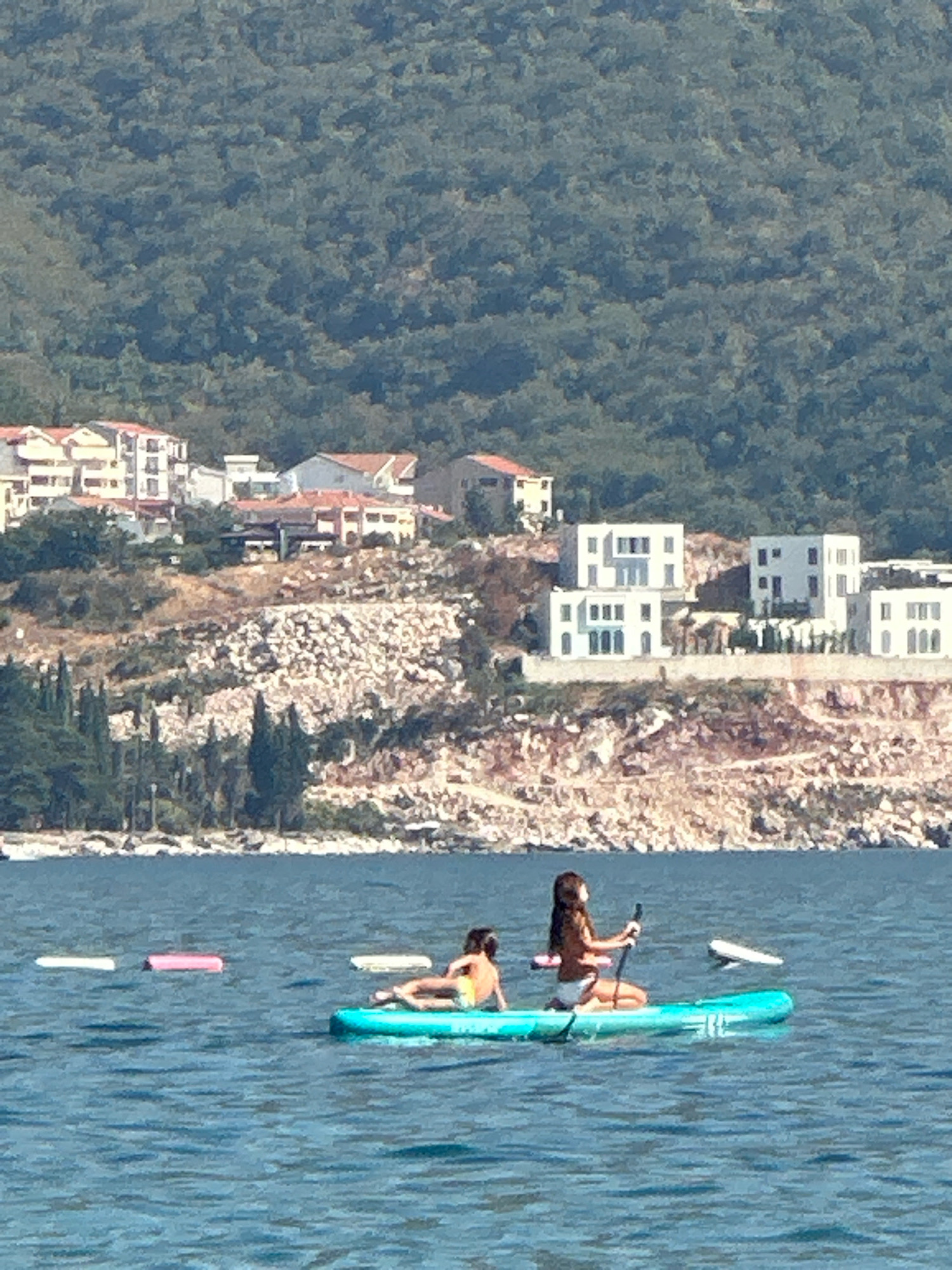 Two people are on a paddleboard in the water. One person is kneeling and holding a paddle, looking towards the right. The other person is lying down on the paddleboard, looking to the left. There are three inflatable floats in the water to the left of the paddleboard. In the background, there is a rocky hillside with several buildings and trees. The buildings are a mix of white and tan, with red-tiled roofs. The hillside is covered in scrub and some trees. The water is a deep blue with small waves. The sky is a light blue and clear. The scene appears to be a sunny day at a coastal location, likely in Boreti, Montenegro, given the architecture and terrain.