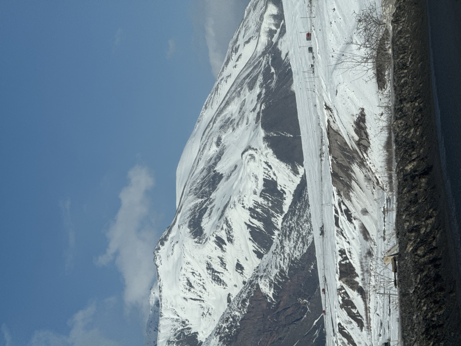 The image depicts a snow-covered mountainous landscape in Kanobi, Georgia, under a partially cloudy blue sky. Snow-covered mountains with exposed rock faces dominate the background. A wide, snow-covered plain with undulating terrain and exposed dark earth occupies the mid-ground. Utility poles and lines traverse this mid-ground. Multiple vehicles are visible on a snow-covered track: two red trucks on the left, and a cluster of lighter-colored vehicles centrally. The immediate foreground consists of a dark asphalt road and a dark, textured embankment. No individuals are discernible.