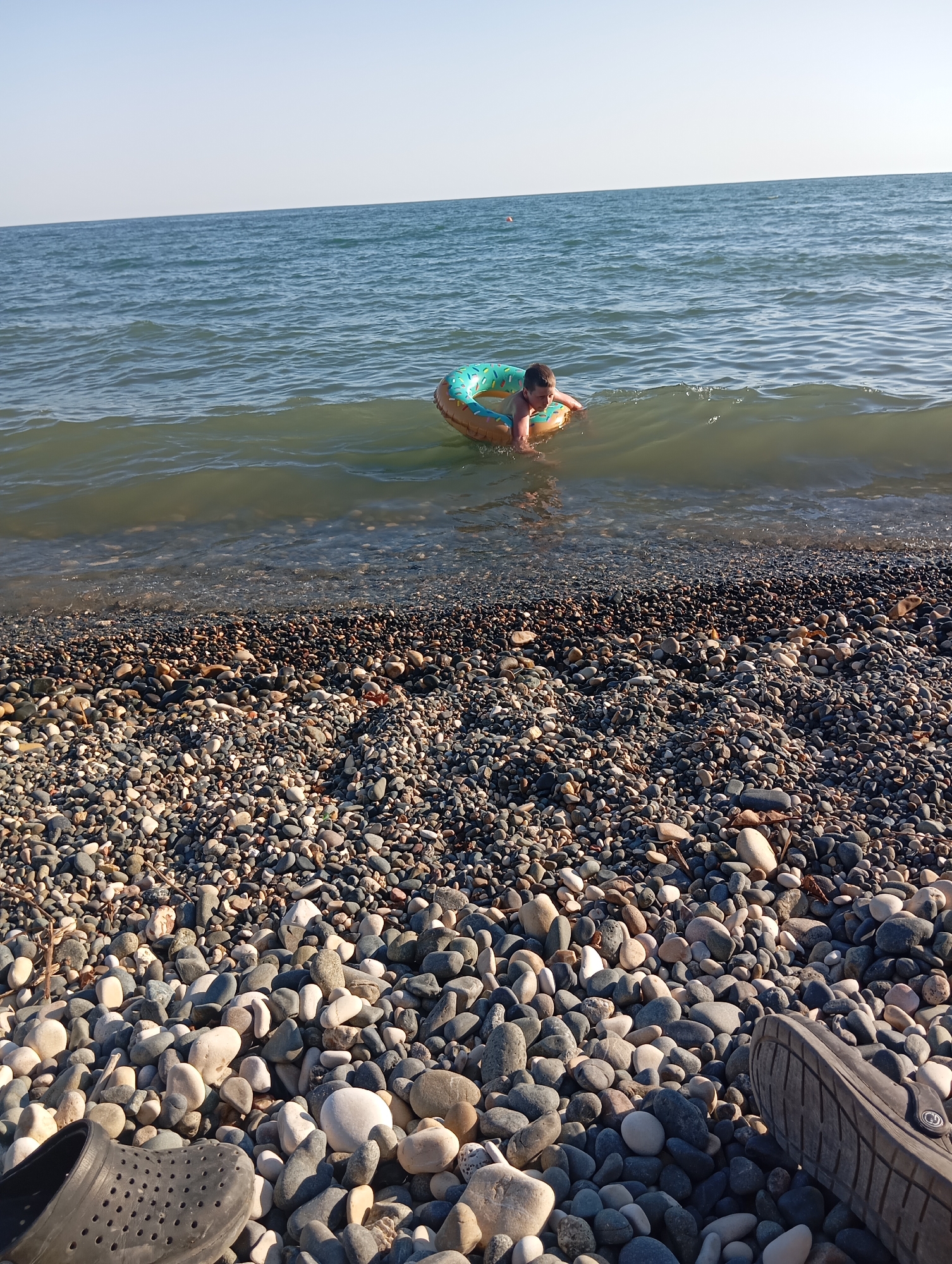 In Sokhumi, Abkhazia, on a sunny day, a young boy floats in the Black Sea on a circular inflatable ring, appearing to be a donut. The water is calm with gentle waves lapping against a pebble beach. The beach is covered in smooth, grey, and white pebbles. In the foreground, a dark-colored clog is visible on the left, and the sole of another dark-colored shoe is partially visible on the right. The sky is clear and blue.