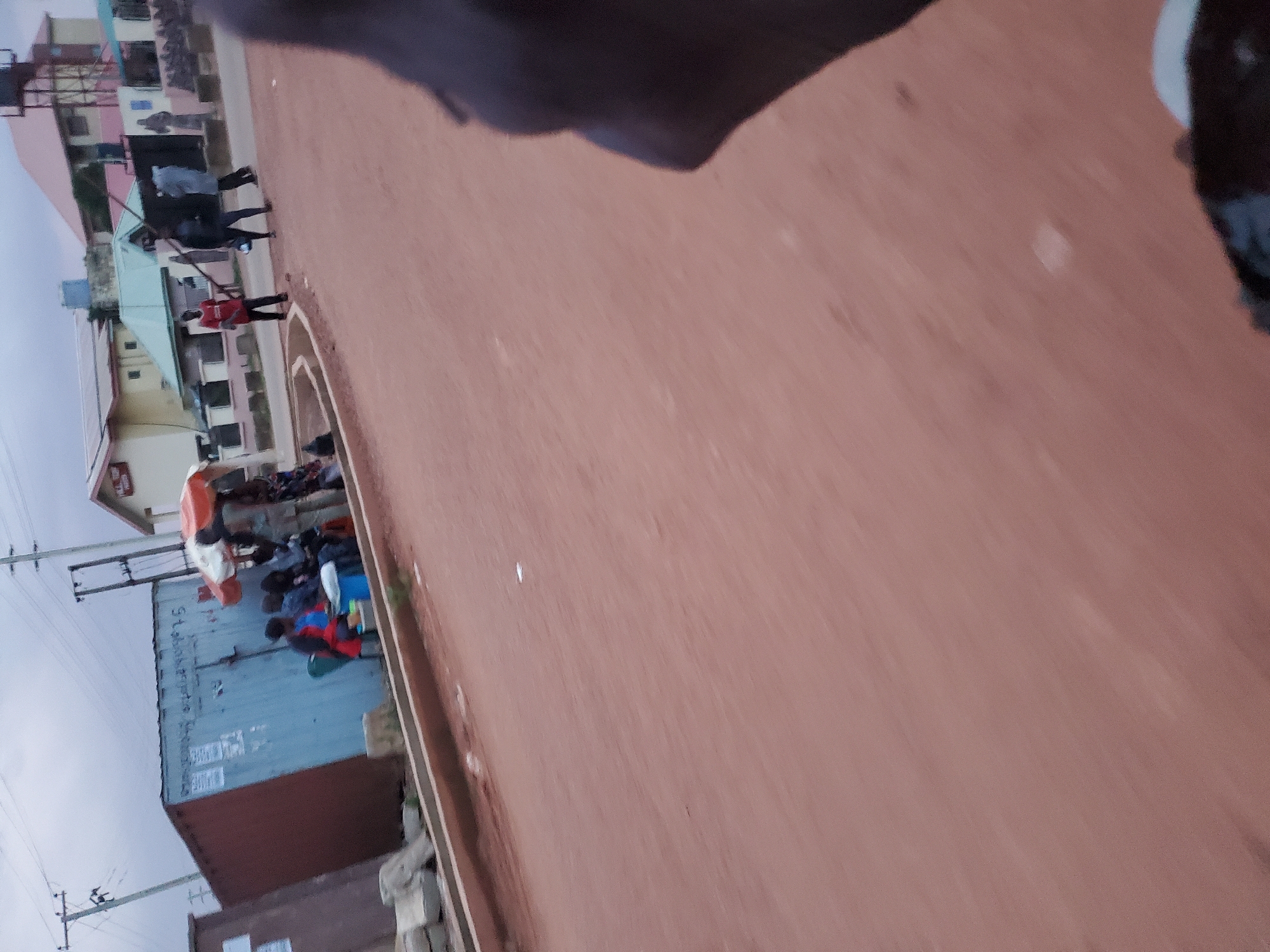 The image depicts a street scene in Gura Top, Nigeria. In the foreground, a portion of a person's dark-skinned hand and arm is visible, suggesting the photo was taken from a moving vehicle. The road surface is reddish-brown dirt.
To the left of the road, a blue shipping container is situated, with some writing and posters visible on its side. A group of people are gathered around the container, some seated and others standing. One individual is wearing a red shirt, and another is wearing a grey garment. An orange and white umbrella provides shade for some of the individuals. To the right of the container, more people are standing on the road.
Further down the road, several buildings with colorful roofs are visible. The sky is overcast. There are utility poles with electrical wires present in the background. The overall environment appears to be a bustling area with everyday activities taking place.