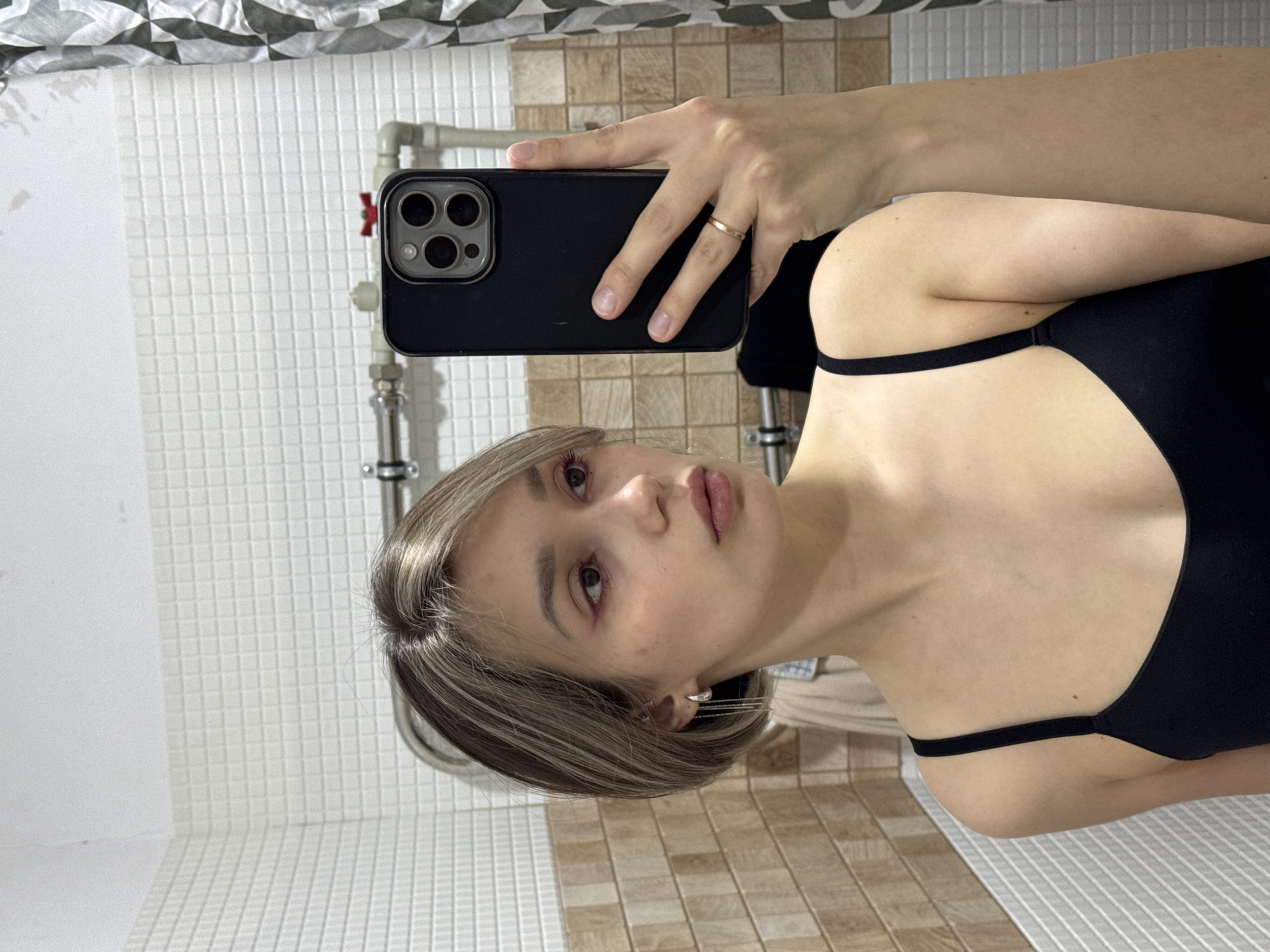 A person is taking a selfie in what appears to be a bathroom. The individual is holding a black smartphone with a triple-lens camera array. The person is wearing a black top with thin straps. The bathroom features tiled walls in varying patterns: white square tiles and beige-brown square tiles. A metal pipe is visible on the wall near the top left of the frame. The location is unspecified.