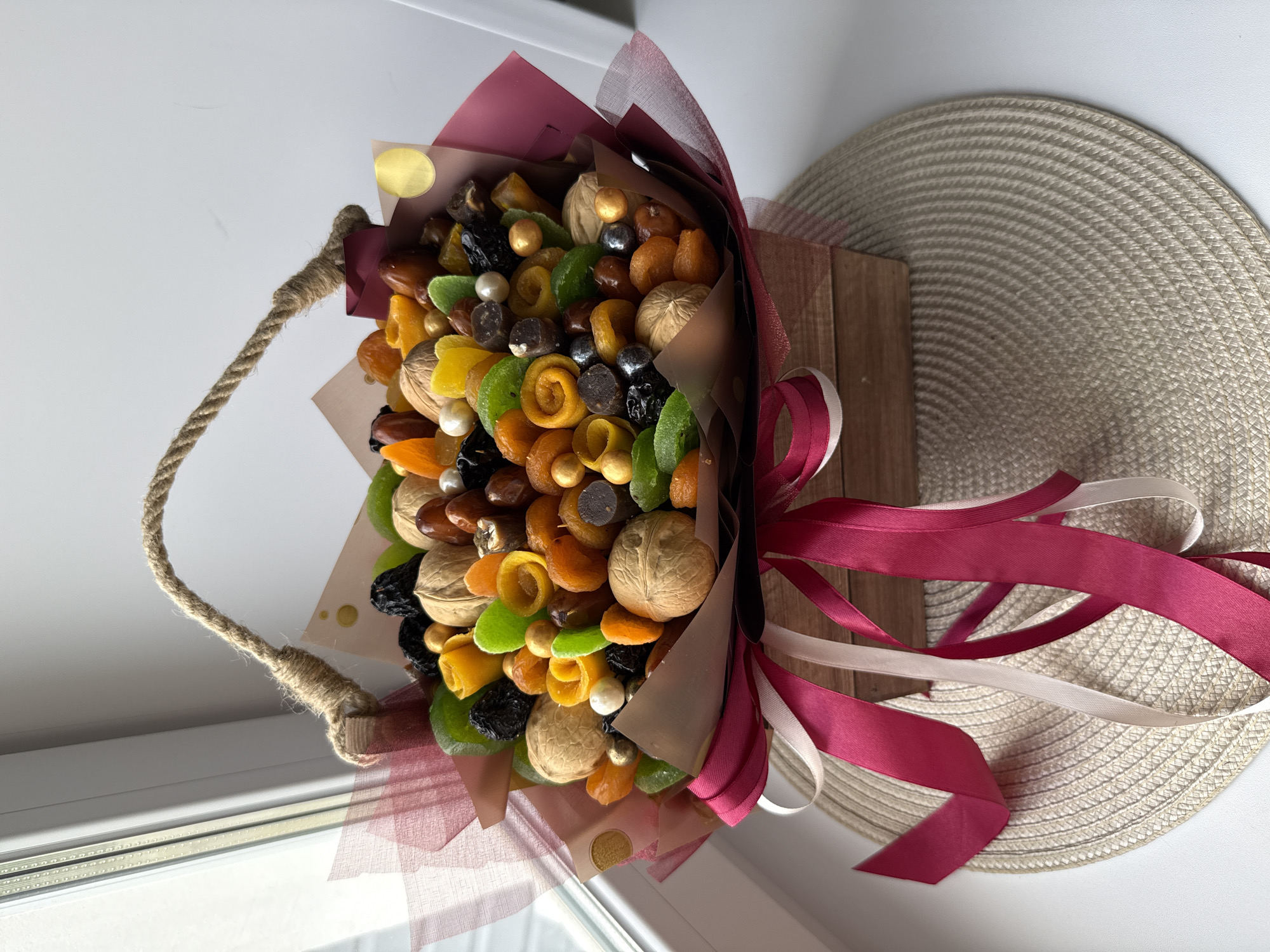 Here is a description of the image:
A bouquet of dried fruits and nuts is presented in a decorative arrangement. The bouquet features a variety of items, including walnuts, dried apricots, plums, and candied fruit pieces in colors like green, yellow, and orange. Small round objects, possibly decorative pearls or nuts, are interspersed among the other items. The bouquet is wrapped in brown and maroon paper, with a pink ribbon tied around the base, and it has a handle made of thick rope. The bouquet is placed on a window sill with natural light coming through. A woven mat is visible in the background. Based on the user prompt, the image location might be in Шебунино, Russia, although the photo does not directly reveal this information.