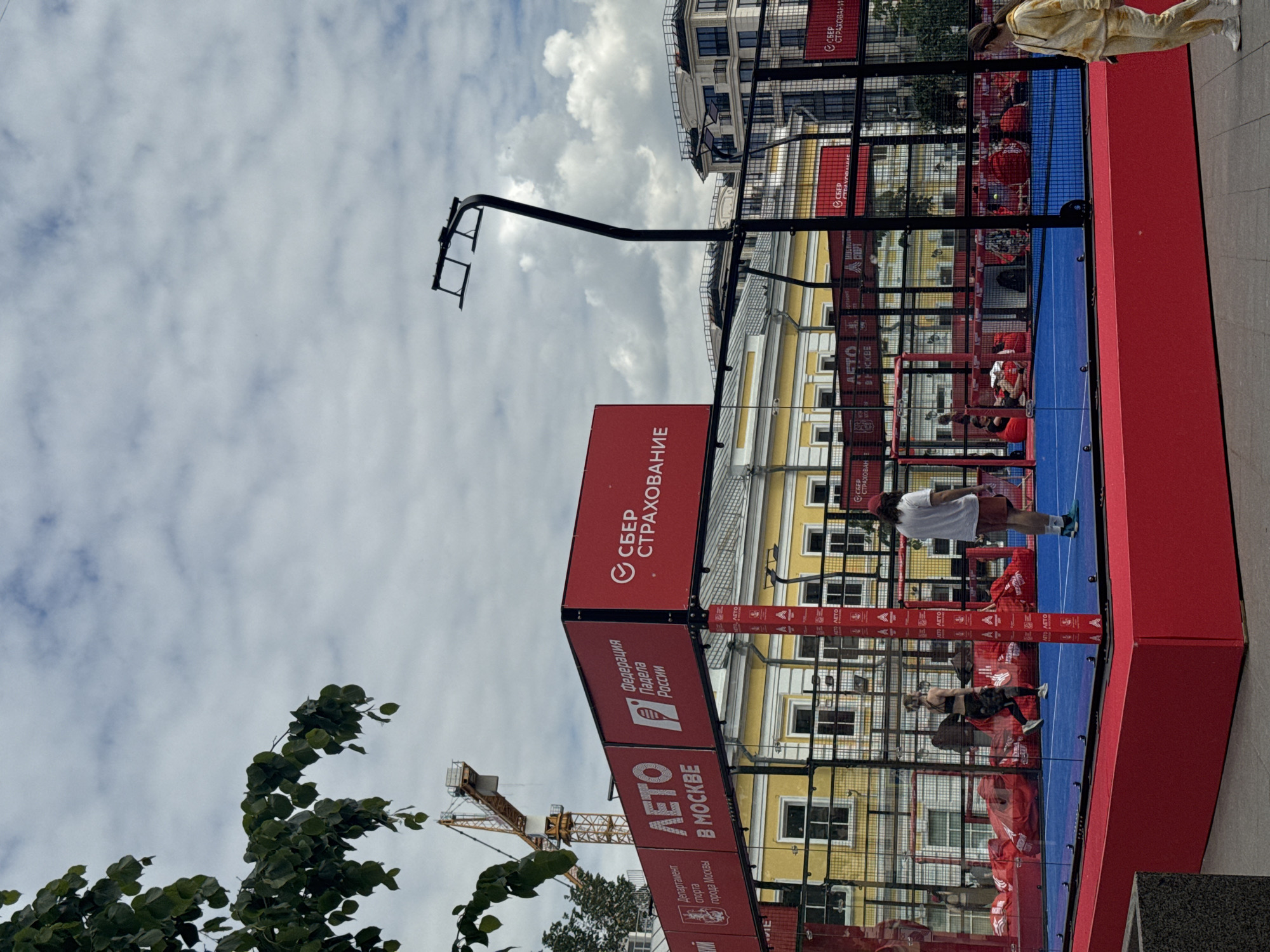 The image, originally rotated 90 degrees counter-clockwise, captures an outdoor padel court situated in an urban setting under a cloudy daytime sky. The court features a vibrant blue playing surface surrounded by red perimeter walls and transparent side panels supported by black metal framing. Several large red banners are prominently displayed on the court's structure.
The banners feature Russian text, including "СБЕР СТРАХОВАНИЕ" (SBER INSURANCE) as a sponsor, "Федерация Падела России" (Padel Federation of Russia), and "ДЕПАРТАМЕНТ СПОРТА ГОРОДА МОСКВЫ" (Department of Sport of the City of Moscow). The phrase "ЛЕТО В МОСКВЕ" (SUMMER IN MOSCOW) suggests a summer sports event or campaign in the city.
Multiple individuals are present. Outside the court, on the right, a person in a light-colored (yellow/beige) tracksuit stands. Inside the court, a person wearing a white t-shirt and dark shorts is visible on the blue playing area, along with another individual in dark athletic wear. Other figures in red are partially seen within the court, possibly spectators or participants.
The background reveals typical city architecture, including a light yellow building with numerous windows and a taller, more modern structure. Trees and a construction crane are also visible, further indicating an urban environment. The scene depicts an active public sports event, likely a padel demonstration or tournament, taking place in Moscow, Russia.
