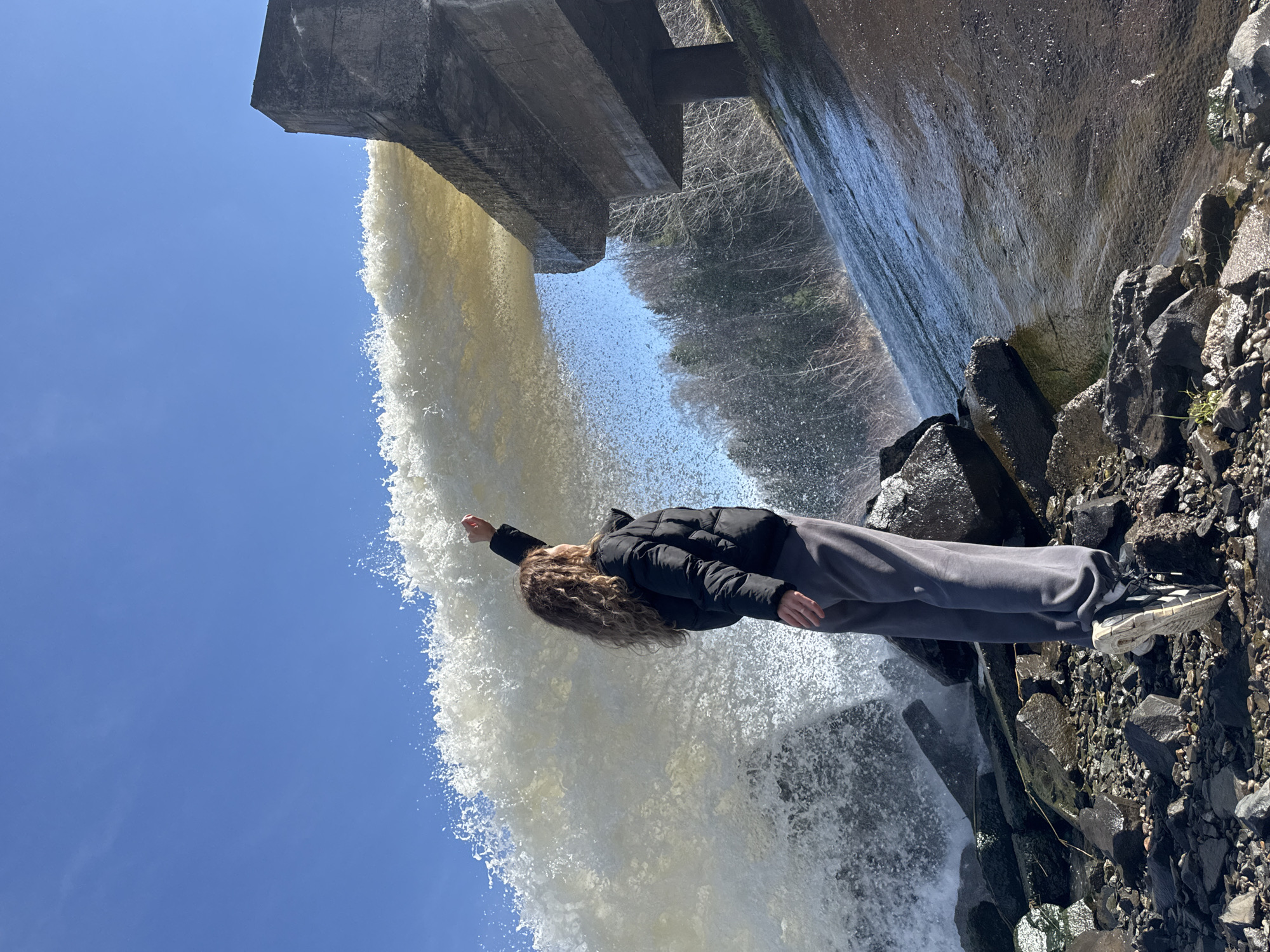 An outdoor, full-length shot depicts a person standing at the base of a powerful waterfall, extending one arm towards the cascading water. The person, viewed from behind and slightly to the side, has long, curly brown hair and is dressed in a black puffer jacket, grey sweatpants, and black and white athletic shoes. The waterfall originates from a large, grey concrete structure, likely a dam or spillway, positioned prominently in the upper portion of the frame. The water is a foamy white and flows with significant force, creating a wide curtain of spray and mist.
The ground around the person consists of dark, wet rocks and gravel. In the background, visible through the water's spray and beyond the concrete structure, are bare trees or a forest, suggesting a late winter or early spring setting. The sky above is a clear, bright blue, indicating a sunny day. The scene captures an adventurous or awe-inspired interaction with the powerful water feature. There is no visible text in the image. The location context is Noviy, Russia.