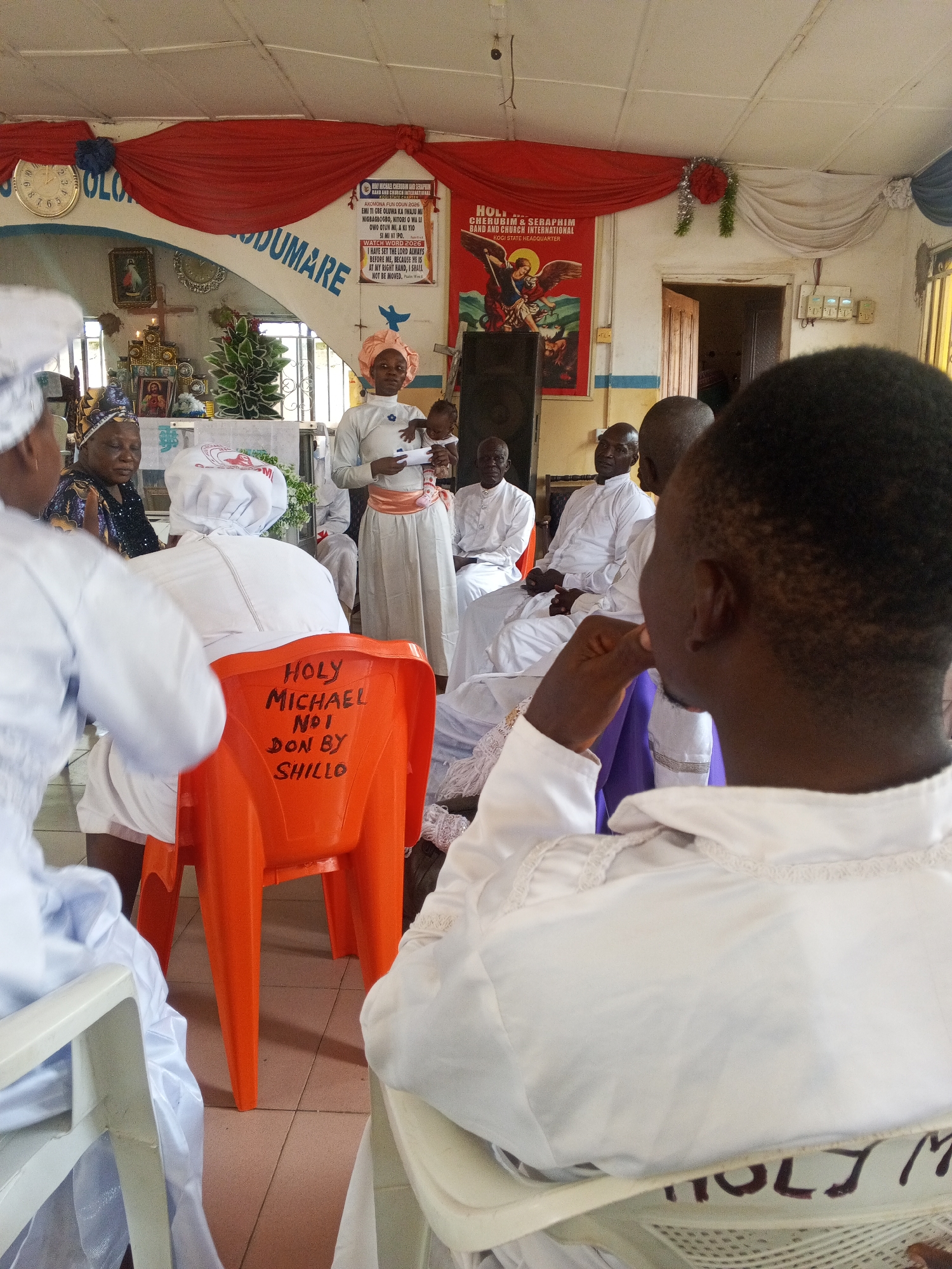 The image depicts an indoor religious gathering at the Holy Cherubim & Seraphim Band and Church International, Kogi State Headquarter, located in Lokoja, Nigeria. The scene shows approximately ten to fifteen individuals, primarily adults, dressed in white ceremonial robes, a common attire within the Cherubim & Seraphim denomination.
In the center, a woman wearing a light-colored dress with a peach sash and headwrap stands, holding an infant dressed in white. She appears to be presenting or speaking, holding a white document or book in her left hand. To her left, an individual is seated on an orange plastic chair, facing away from the camera. The back of this chair has "HOLY MICHAEL NO1 DON BY SHILLO" handwritten in black capital letters.
The background features the church's interior architecture and decorations. A prominent red and white fabric drape spans the upper wall section, adorned with a silver and red tinsel decoration on the right. Below this, an arched architectural feature is inscribed with text, part of which reads "OLODUMARE". On the left wall, a gold-colored analog clock displays a time past 12.
Two large posters are visible on the back wall. The left poster is predominantly red and white, stating "HOLY CHERUBIM & SERAPHIM BAND AND CHURCH INTERNATIONAL KOGI STATE HEADQUARTER" and includes text in what appears to be Yoruba, along with a Bible verse reference (Psalm 16:8). The right poster, also red, depicts a winged figure, presumably Archangel Michael, slaying a dragon-like creature, with the same church name and "KOGI STATE HEADQUARTER" printed.
To the left of the central woman, an altar or shrine-like area is visible, featuring a cross, lit candles, framed religious images (including one of Jesus and one of Mary), and a small green decorative plant. A dark speaker is positioned near the left wall. Several men in white robes are seated in the background, observing the proceedings. The floor is tiled with light-colored square tiles. A slightly ajar wooden door and a window are visible on the right side of the background. Electrical switches are mounted on the wall near the door. The ceiling is white with visible structural elements and possibly light fixtures.