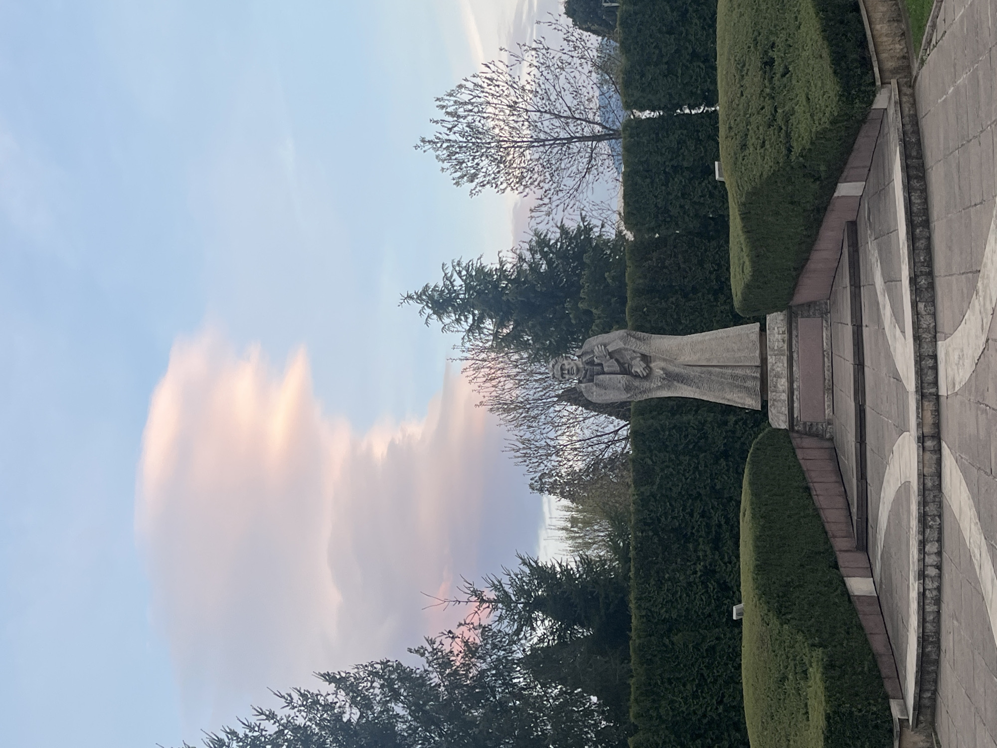 This image presents an outdoor scene dominated by a monumental stone sculpture within a formally landscaped environment, likely a park or memorial in Sofia, Bulgaria. The central subject is a light-colored stone sculpture of a human figure, positioned on a tiered base to the right. The figure, possibly male, is depicted with detailed clothing or drapery and a composed expression, appearing to gaze towards the left.
The sculpture is flanked by neatly manicured, dark green hedges that create structured lines and paths throughout the midground. Beyond these hedges, various trees contribute to the setting; some show intricate, bare branches against the sky, suggesting a season like late autumn or early spring, while others are dense evergreens.
The ground in the foreground and extending along the right side consists of a paved area or walkway. This paving features a distinctive pattern of light and dark gray stone tiles arranged in interlocking arcs or segments, adding to the formal design of the space.
The sky occupies a significant portion of the background, characterized by dramatic, elongated clouds. These clouds are illuminated with soft, warm hues of pink and orange, particularly on the left, suggesting the golden hour of either sunrise or sunset. The overall lighting is gentle and diffused. The scene evokes a sense of solemnity and peace. No activity is depicted, and no living individuals are visible. There is no visible text in the image.