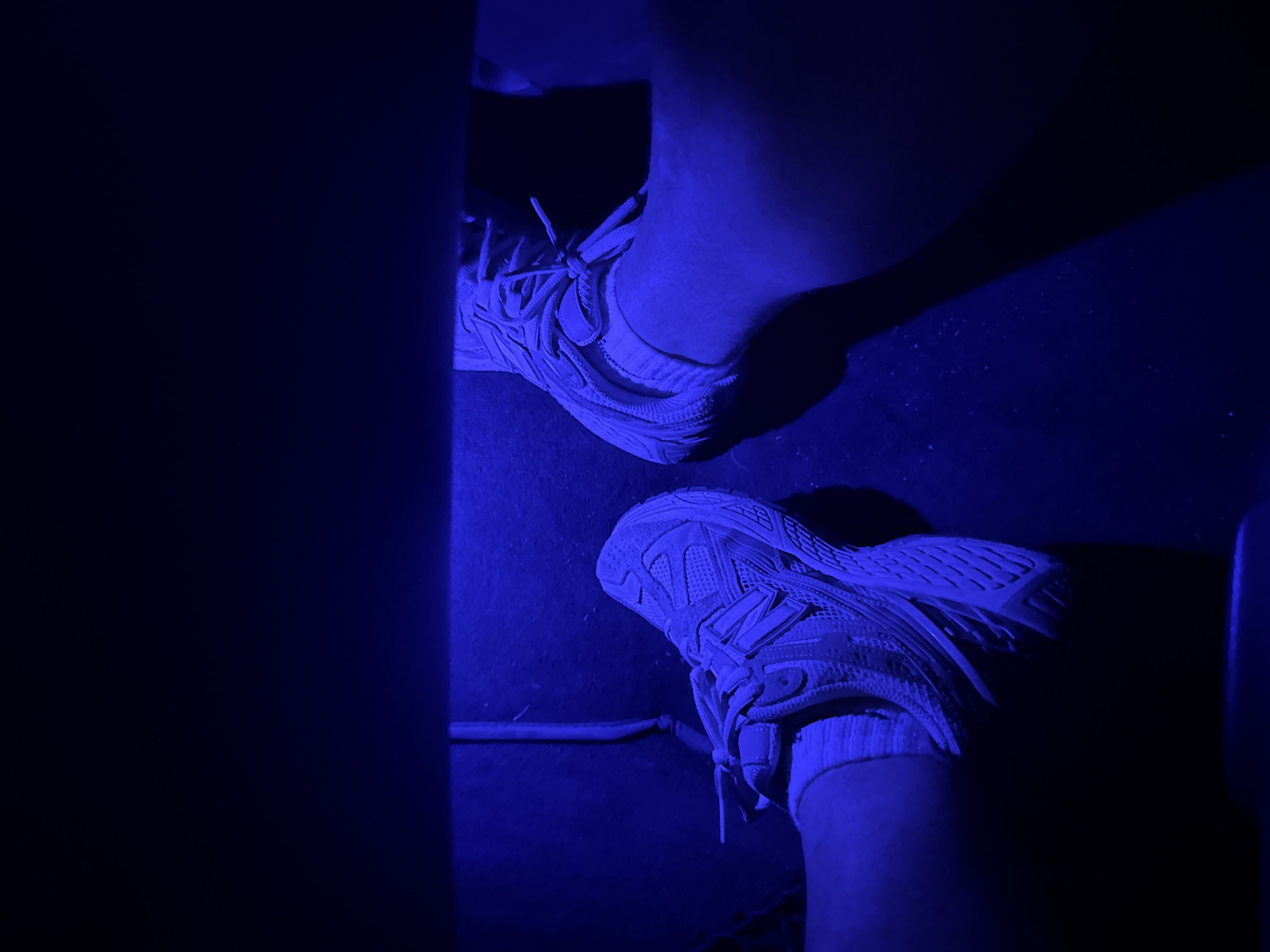 The image presents a close-up, low-angle view of two feet wearing white sneakers, likely New Balance brand, situated on a dark surface. The scene is predominantly illuminated by a blue light source, casting strong shadows and altering the colors. One foot is positioned slightly above the other in the frame. A thin, dark rod or bar is visible near the lower foot. A dark, undefined object obscures the left side of the image, potentially a wall or piece of furniture. The environment appears to be indoors. There are no people or specific actions visible other than the presence of the feet and shoes. The specific location in Rajana, Bulgaria is not ascertainable from the provided image.