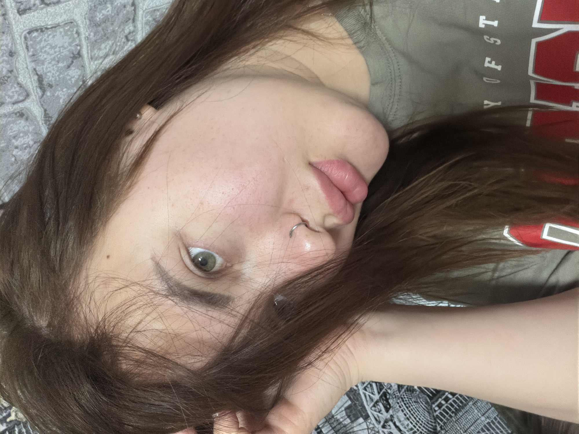 A young woman with brown hair and green eyes is lying down. She is wearing a gray-green shirt with red and white lettering that reads "TY OF ST." She has a nose ring and a piercing in her ear. The background includes a gray, textured surface.