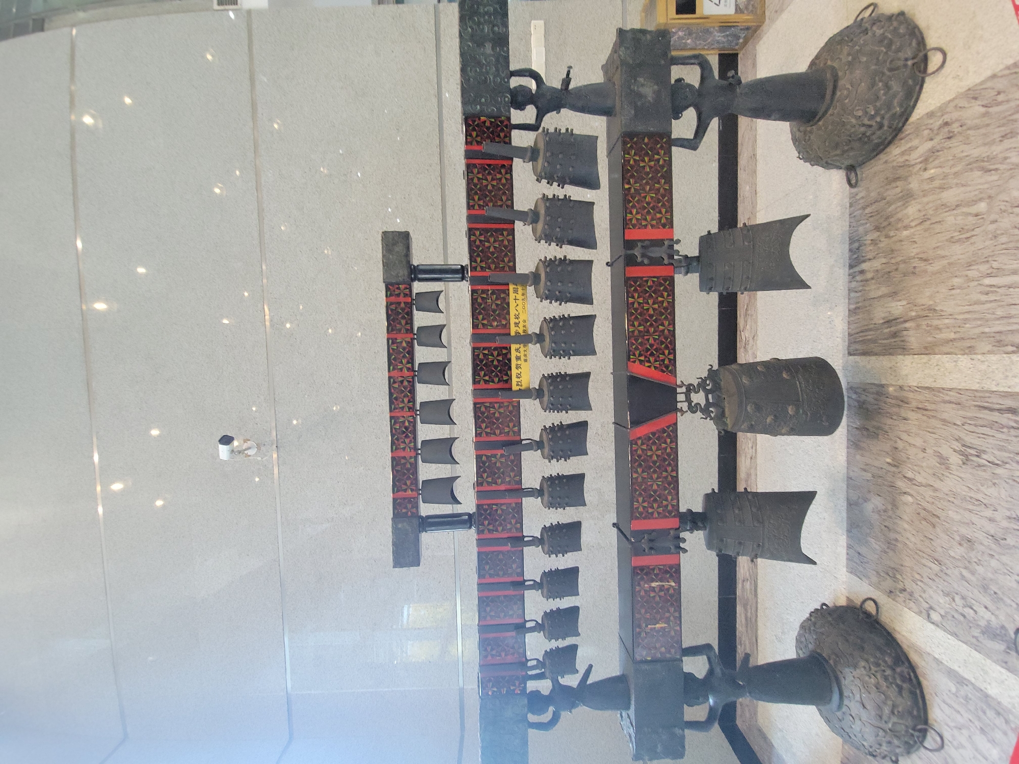 The image showcases a set of ancient Chinese chime bells displayed in a museum. These bells, arranged in three rows, are of various sizes and shapes. The supporting structure is made of dark-colored wood with intricate red and gold patterns. The bells themselves are made of a dark metal, likely bronze or iron.
The environment appears to be a modern museum space, as indicated by the polished stone or tile flooring and the smooth, pale-colored wall. The presence of artificial lighting is also evident, casting reflections on the floor and walls.
A small plaque with Chinese characters can be seen on one of the columns. In the background, there is a security camera on the wall.
The museum's location is identified as Shapingba District, China, based on the Chinese characters on the plaque, which mentions Chongqing University.