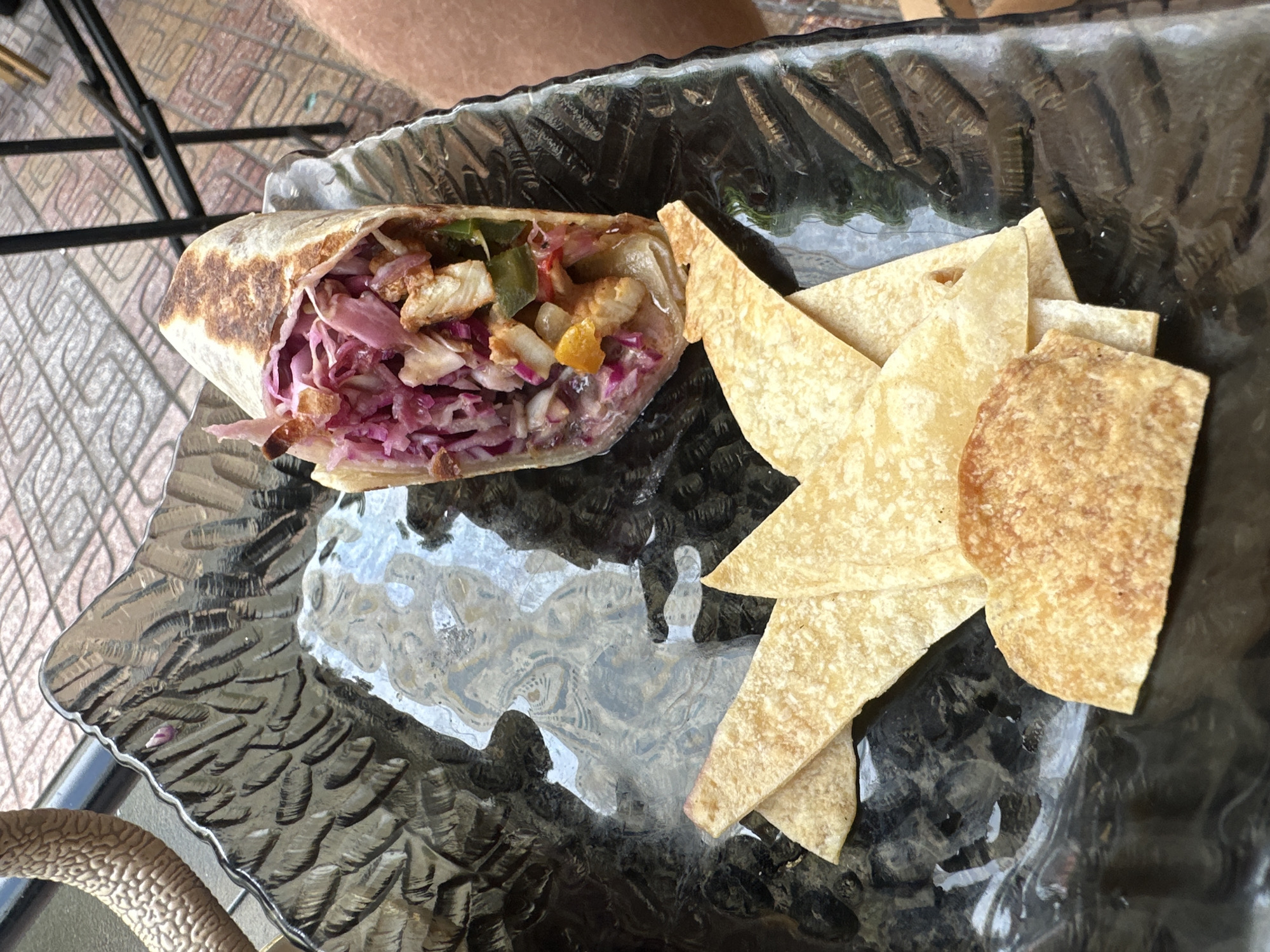 A close-up, overhead shot shows a street food dish. The dish contains a fully loaded burrito on a dark, textured plate. The burrito appears to be filled with chicken, red cabbage, and possibly other vegetables. To the left of the burrito are several pieces of tortilla chips, some triangular and some rounded. The plate is set on a patio with a patterned tile floor. The background is out of focus, but appears to be an outdoor seating area with a table leg visible. The lighting suggests it's daytime.