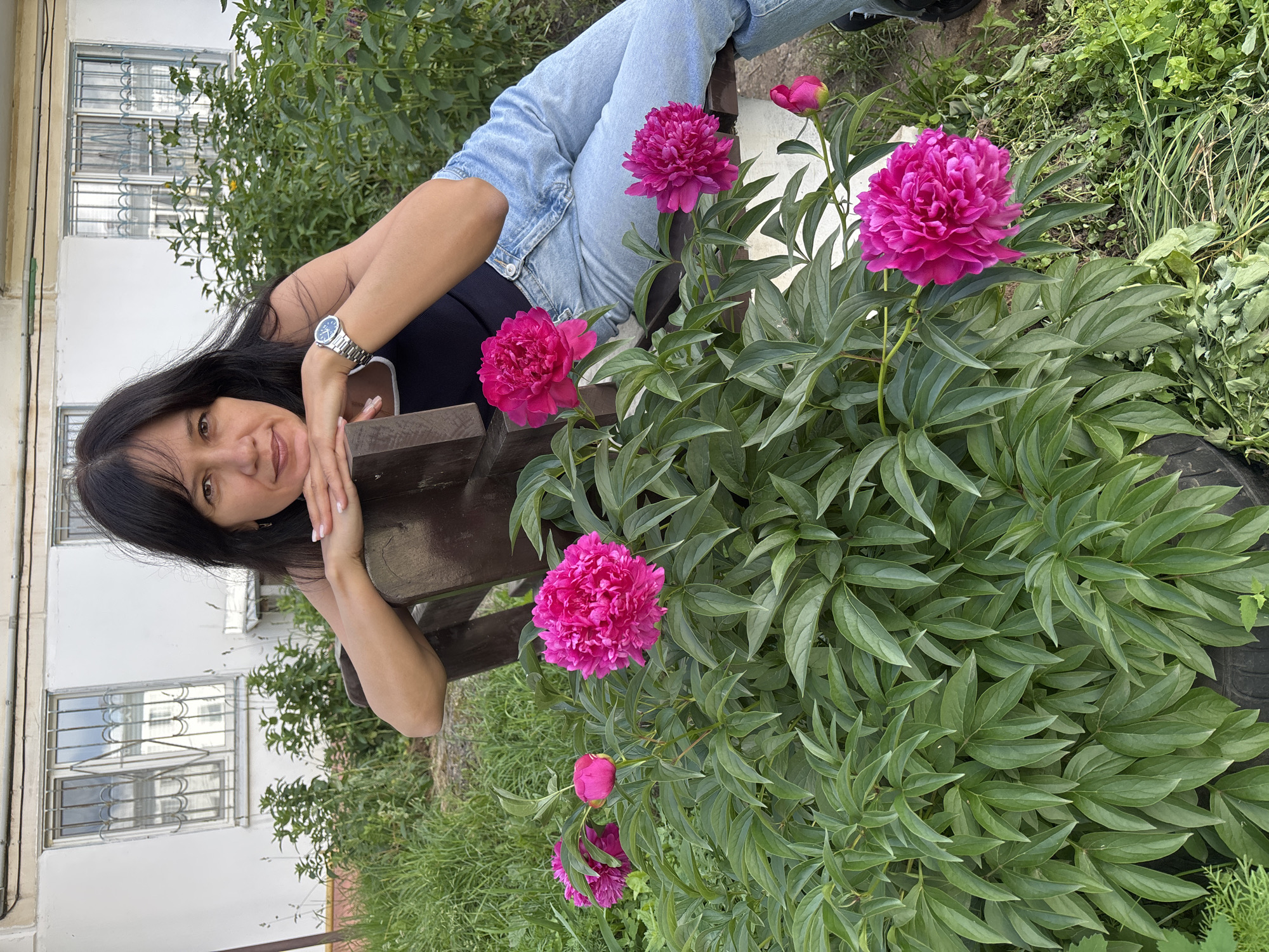 A woman with long dark hair is leaning on a wooden bench, resting her chin on her hands. She is wearing a black top and light blue jeans. She is looking at the camera and smiling. In front of her, a bush of bright pink peonies is in bloom. The background shows a building with white walls and windows, surrounded by greenery. The scene appears to be outdoors during the daytime. The overall mood is relaxed and pleasant. There is no visible text in the image.