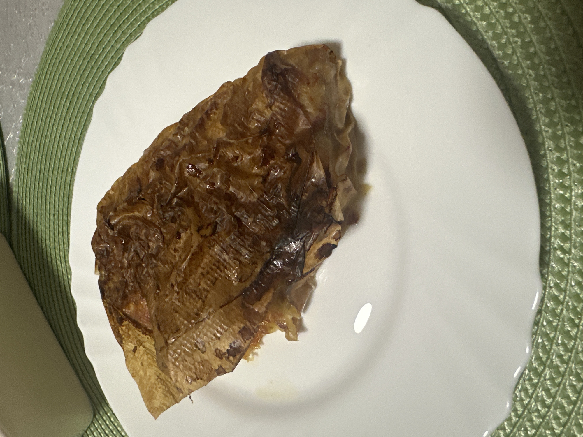 The image shows a piece of cooked food, possibly tofu skin, sitting on a white plate. The plate is placed on a green placemat, which is on a textured, possibly gray or white, surface. The location in the city of Vorotynsk, Russia is unknown and cannot be derived from this image. There are no people visible in the frame.