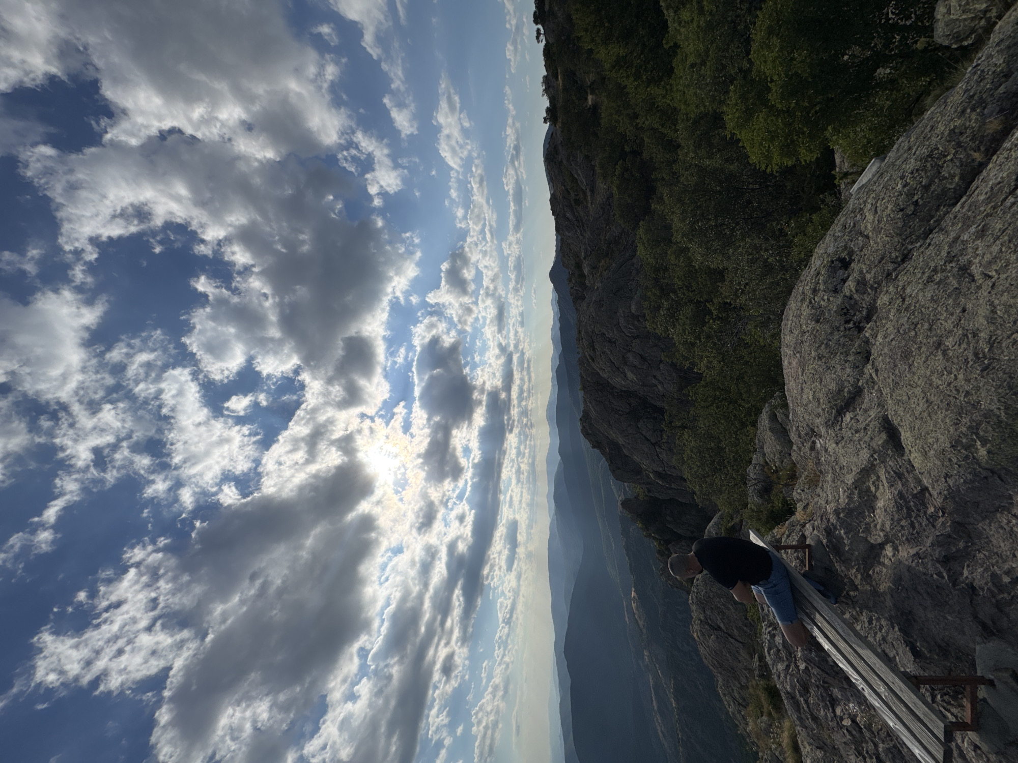 A man sits on a wooden bench overlooking a vast mountain landscape. The scene is captured from a high vantage point, with a dramatic drop into a valley of layered, hazy mountains stretching into the distance. The sky is filled with a mix of white and grey clouds, with the sun peeking through, creating shafts of light. The man is dressed in a dark t-shirt and blue shorts, with his back to the camera, leaning forward as if contemplating the view. He is bald and appears to be looking down. The foreground is dominated by a rough, grey rock face and the wooden bench, which is set against the rocky terrain. To the right of the frame, lush green trees cling to the side of the mountain. The lighting suggests it is daytime, possibly late afternoon or early morning due to the hazy distant mountains and the angle of the sun. The overall mood is one of quiet reflection and awe inspired by the natural scenery. The location is Ichera, Bulgaria. No visible text is present in the image.