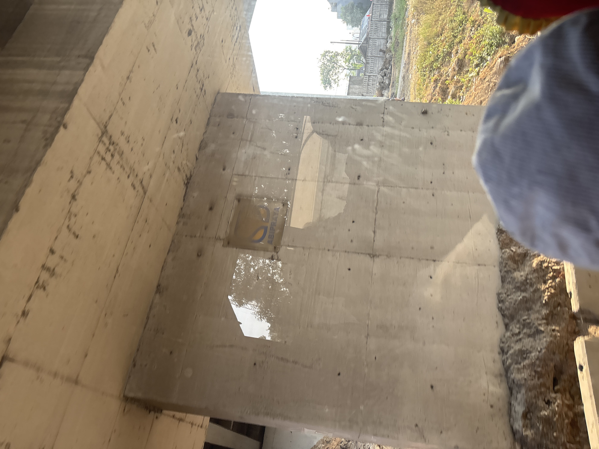 The image depicts a concrete pillar supporting an elevated structure, likely a bridge or overpass, in Jayamukti, Indonesia. The pillar has a reflective surface, showing a reflection of trees and the sky. A small yellow sign with blue lettering is affixed to the pillar. The surrounding environment includes dirt, vegetation, and a concrete fence in the background. The perspective suggests the photo was taken from a moving vehicle or from a ground-level position looking up and to the side. A person's head, wearing a grey cap, is visible in the foreground, partially obscuring the view of the pillar.