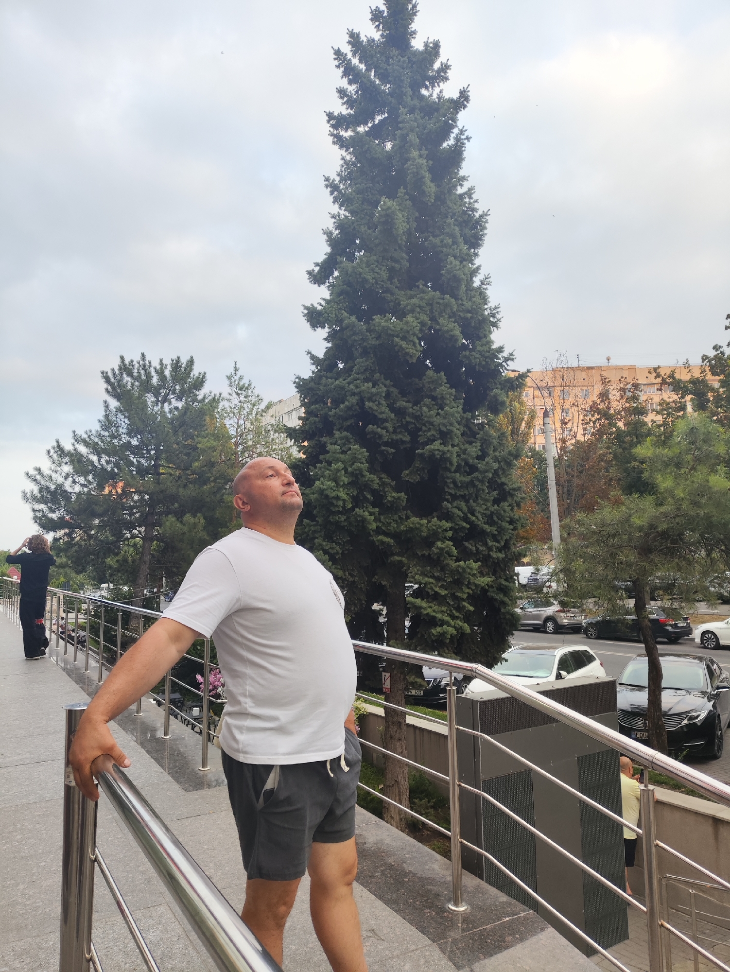 A man with a shaved head is standing on a paved pathway with a metal railing on the right. He is wearing a white t-shirt and gray shorts and is looking upwards with his eyes closed. Behind him, another person in a black outfit with their back to the camera is walking away. Tall evergreen trees and buildings can be seen in the background, with cars parked along a street. The sky is overcast. This scene appears to be from Chișinău, Moldova.