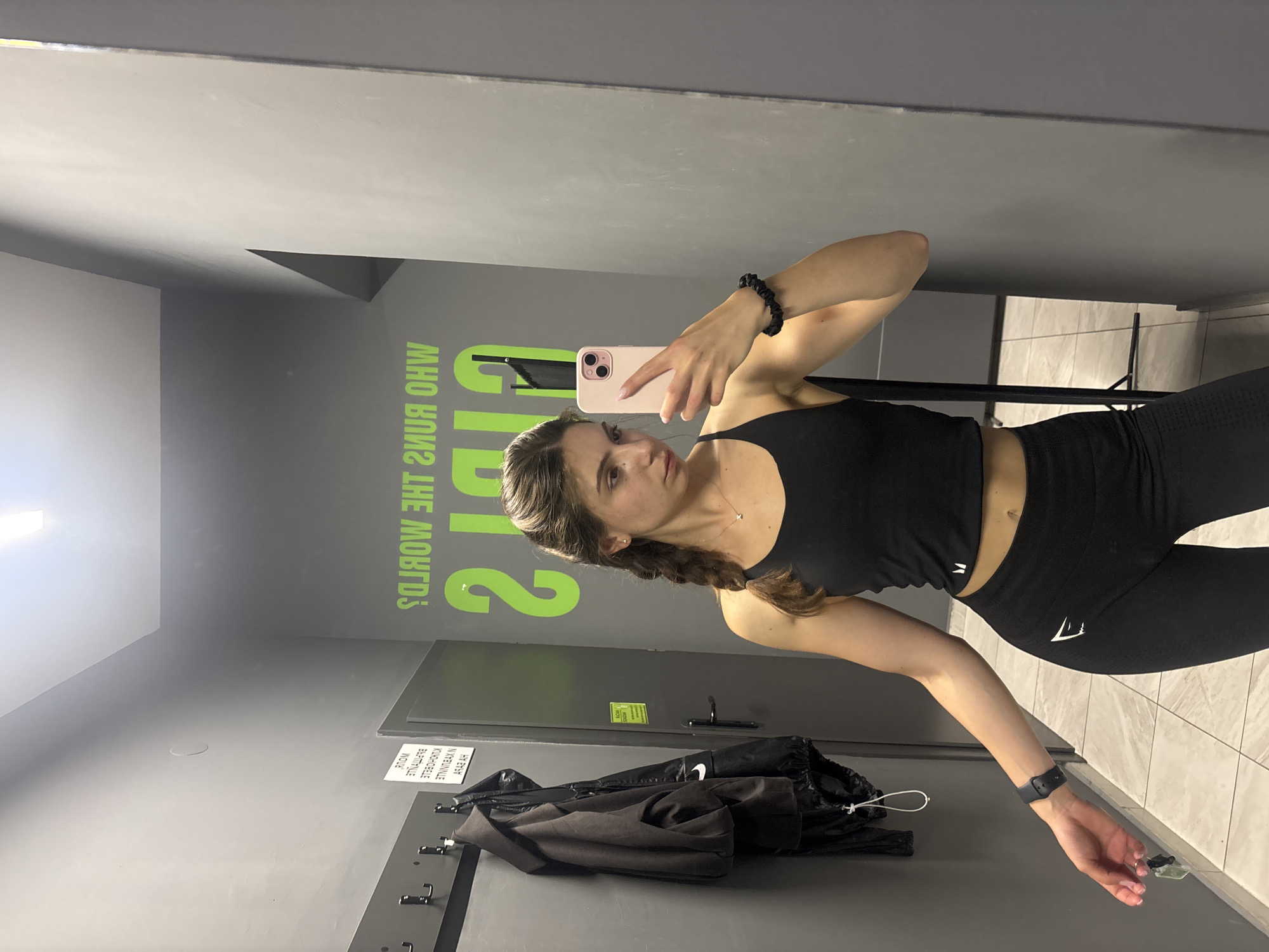 The media file is a mirrored selfie taken by a young woman in what appears to be a gym changing room or locker room in Varna, Bulgaria.
The subject is a woman with brown hair, styled in braids, wearing a black sports bra/crop top and matching black leggings with a visible white Gymshark logo. She has a black scrunchie on her right wrist and a black smartwatch on her left. She is holding a rose gold/pink iPhone to take the photo, looking towards the camera in the mirror with a neutral expression.
The setting features two-toned walls: a darker gray on the lower section and a lighter gray above, with a lighter gray ceiling. The floor is tiled with light-colored, possibly cream or light gray, rectangular tiles. In the background, visible through the mirror's reflection, there are coat hooks on the wall, from which a black jacket and a black drawstring bag are hanging. A closed gray door is also visible.
A prominent feature is the bright green text on the wall, mirrored and inverted due to the reflection, which reads "WHO RUNS THE WORLDS?". A large stylized "2" is integrated into this phrase. Below the hooks, a white rectangular sign, also mirrored and inverted, displays text in Bulgarian that translates to "NO PROHIBITED ITEMS IN THE CHANGING ROOM". This confirms the location context of Varna, Bulgaria. The lighting appears to be artificial, typical of an indoor facility.