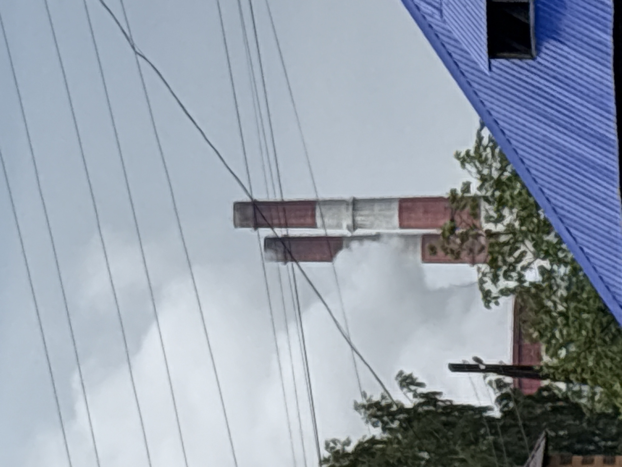 The image shows a view of industrial smokestacks with smoke emitting from them, against a cloudy sky. Multiple power lines are visible crisscrossing the frame. In the foreground, a blue, corrugated metal roof of a building is prominent, with a small, dark-framed window. Trees with green foliage are also present, partially obscuring the view of the smokestacks and some buildings. The scene suggests an industrial area with residential elements in Yayva, Russia.
