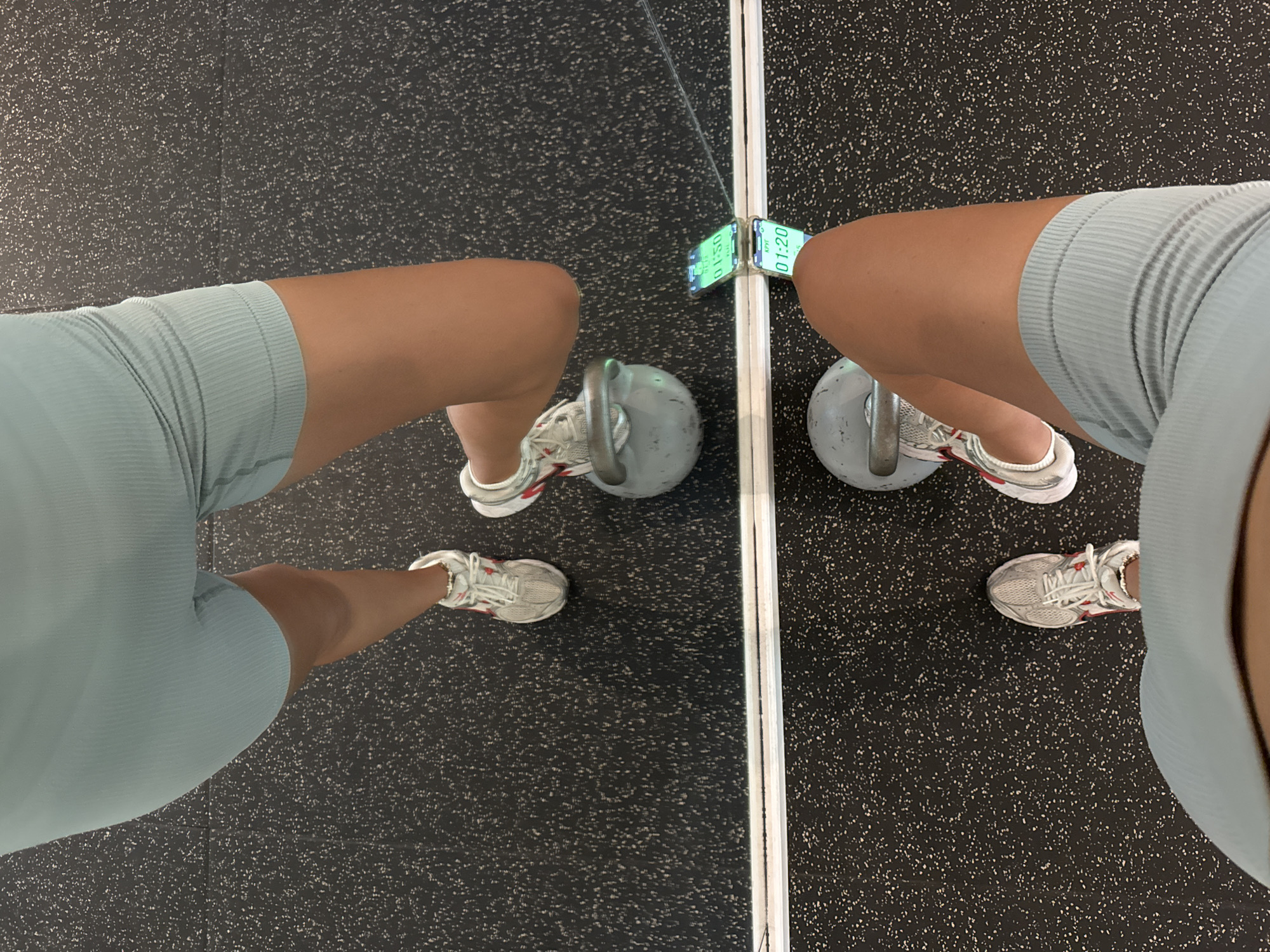 A person is seen from an overhead perspective in an indoor gym setting in Kuta Selatan, Indonesia. The person is wearing light blue athletic shorts and white running shoes with red accents, standing on a black rubber gym floor speckled with white dots. A gray kettlebell is positioned between their feet. A mirror in front of the person reflects the scene, including a smartphone attached to the mirror with a green screen displaying a time.
