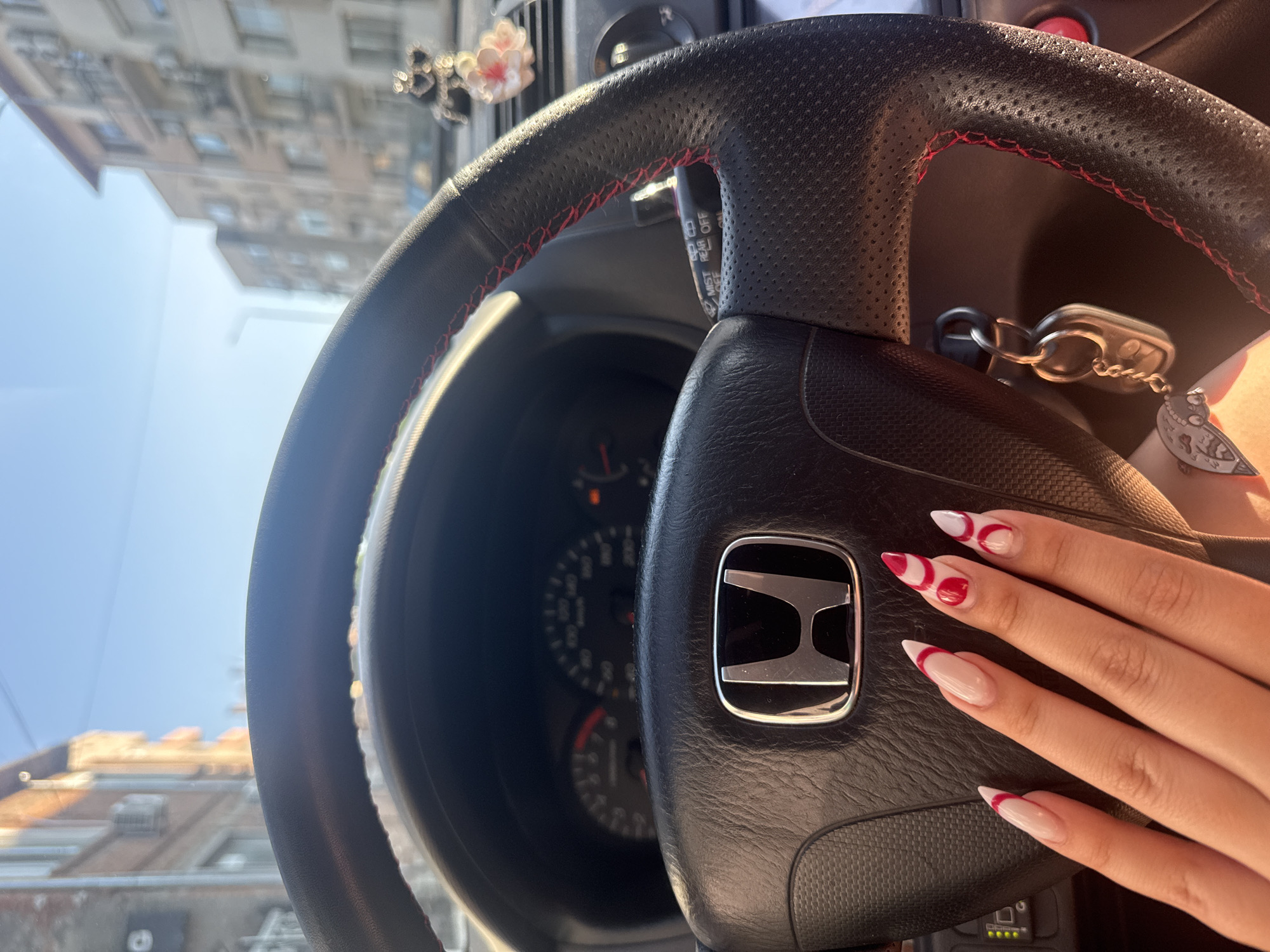 The image shows the interior of a car, focusing on the steering wheel with a Honda logo. A hand with elaborately designed nails rests on the steering wheel. The car keys dangle from the ignition. Through the windshield, a building is visible, hinting at an urban environment. Although the image was purportedly taken in Elhovo, Bulgaria, there are no details that confirm this location.
