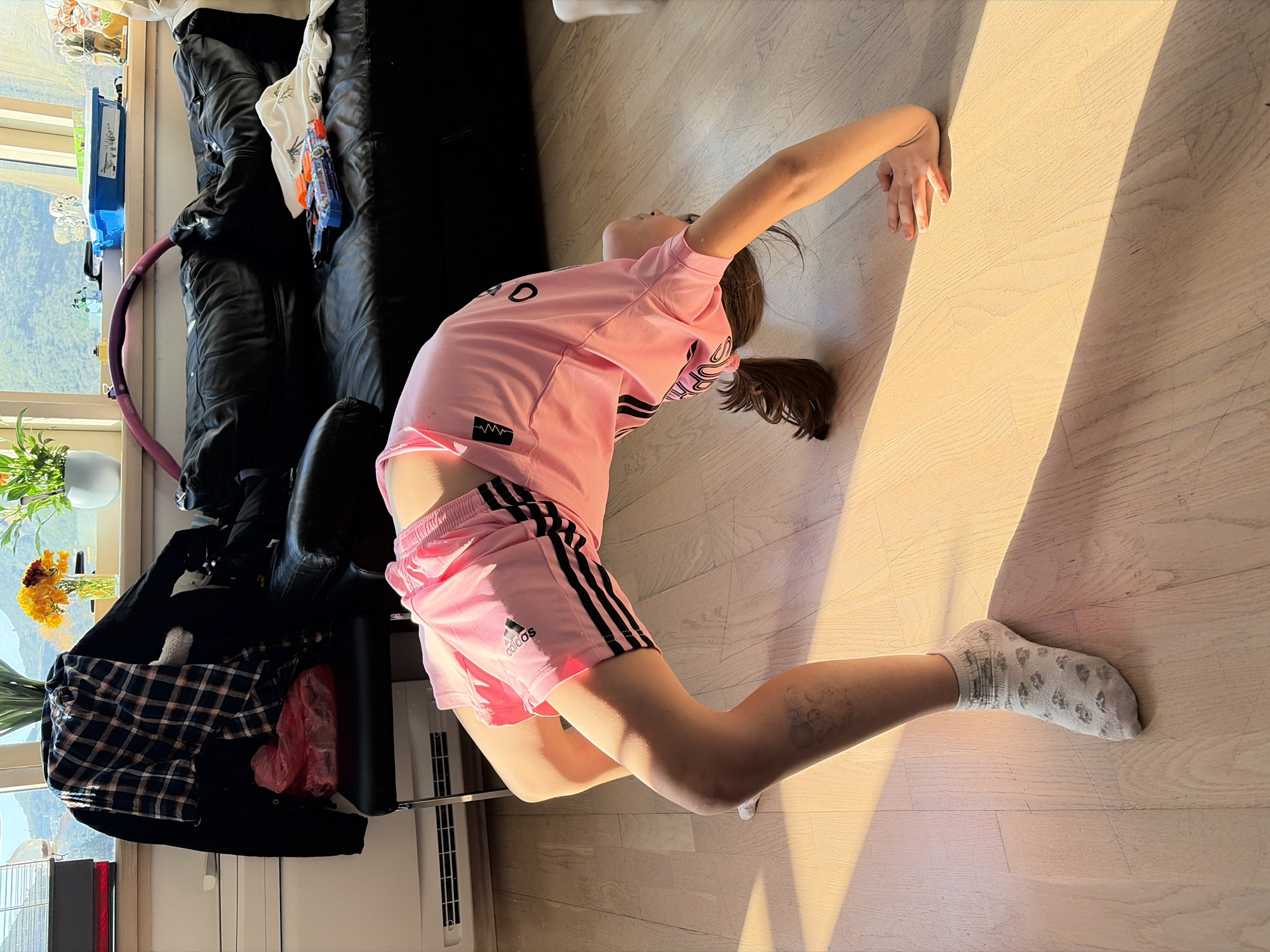 A girl in a pink Adidas outfit with black stripes is performing a backbend on a light wooden floor. She is wearing a white sock on her right foot. The environment includes a black leather couch with a hula hoop, a window with a view of a landscape, a plant with yellow flowers, and a black chair with clothes draped on it. The location appears to be indoors, possibly a residence in Budva, Montenegro, given the landscape visible through the window.