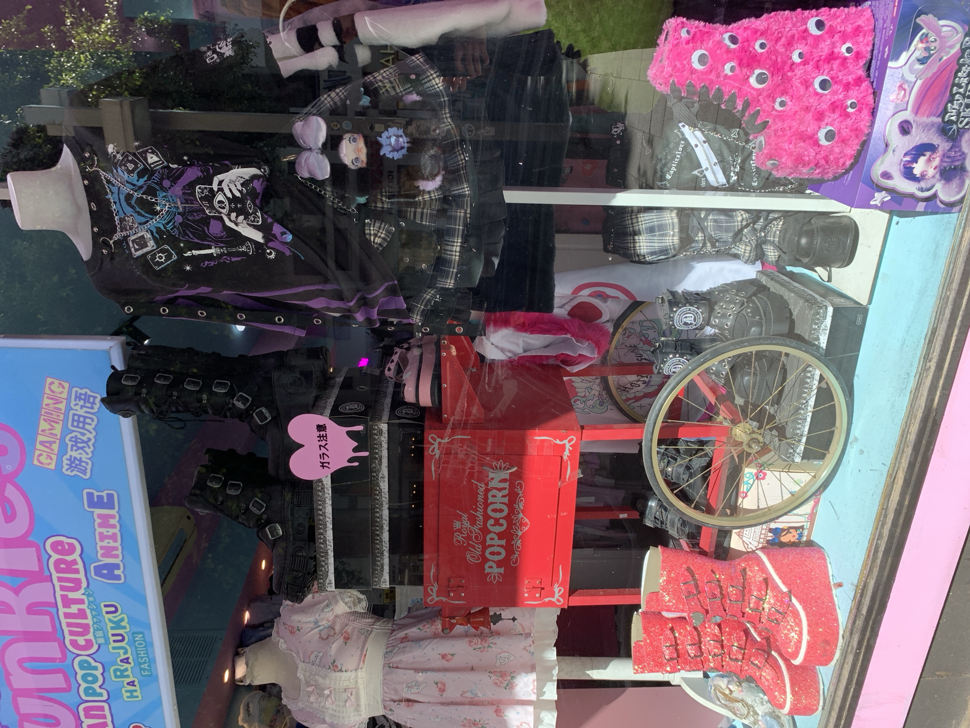 The image shows a storefront window in Tel Aviv, Israel, displaying a variety of fashion items and related objects. The sign next to the window states “HARAJUKU ANIME” and POP CULTURE (referencing Harajuku district). Items displayed include clothing on mannequins (e.g., a pink dress with floral prints), several pairs of distinctive shoes and boots (e.g., buckled boots, red glitter boots), a popcorn machine painted red, and accessories like a pink fuzzy bag with eyes and plaid bags. There is also a sign that says "ガラス注意" (Glass Caution). The reflection of street outside can be seen in the window.