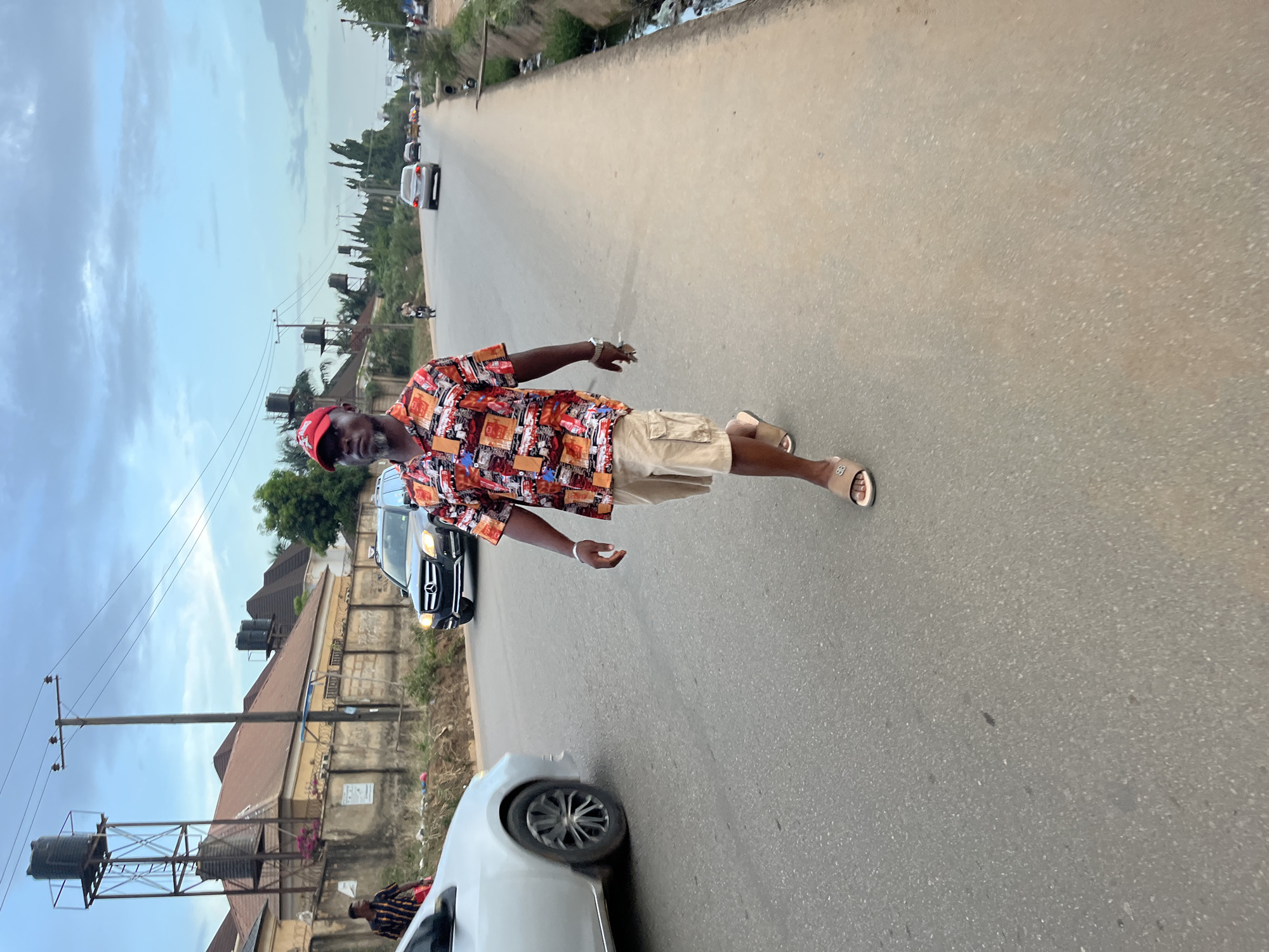 A dark-skinned man stands prominently in the middle of a paved road, posing for a full-body shot in an urban or semi-urban setting in Abuja, Nigeria. He is centrally located in the frame, looking directly at the camera with a neutral expression.
The man is wearing a red baseball cap, a vibrant, short-sleeved button-up shirt patterned with what appears to be cars and buildings in various colors, light-colored (khaki) cargo shorts, a silver-toned watch on his left wrist, and light-colored slip-on sandals. His right hand is slightly open by his side, while his left arm is also relaxed.
The road is light grey asphalt and stretches into the distance, with several vehicles visible. In the immediate foreground, a white sedan is partially visible on the left. Further down the road, a black sedan is parked on the left side, and a silver sedan is on the right. A person on a motorcycle is also seen in the distance on the right. The sides of the road are lined with buildings, fences, and utility poles with wires. Several black plastic water tanks on metal stands are visible near some of the structures, along with scattered trees and vegetation.
The sky is overcast with diffuse clouds, indicating either an overcast day or perhaps late afternoon lighting. The scene suggests a casual moment on a residential or mixed-use street. No visible text is discernable in the image.