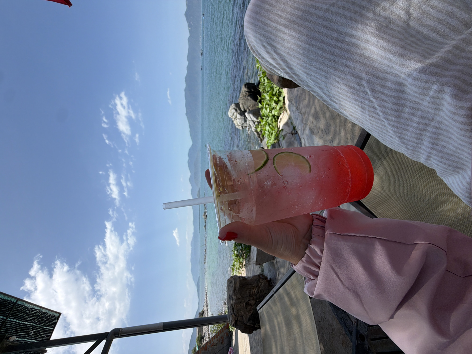 The image displays a first-person perspective of a coastal scene in Vĩnh Lương, Vietnam. A person is seated, holding a clear plastic cup filled with a pink beverage, ice, and two lime slices, with a clear plastic straw inserted. The hand holding the cup exhibits red nail polish on the thumb and index finger. The person is wearing a light pink long-sleeved top with ruched cuffs and light-colored, vertically striped pants.
In the immediate foreground, part of a lounge chair or similar outdoor seating is visible on the left. The ground consists of light-colored concrete or paving, transitioning to natural rocks and green ground cover near the water's edge. To the left, a dark metal support structure for an overhead canopy or awning is present, with a textured dark material partially visible above.
The mid-ground features a calm body of blue-green water, indicative of an ocean or bay, with a rocky shoreline. Several small, dark vessels are visible on the distant water. In the far background, a range of hazy, blue-tinted mountains extends across the horizon. The sky is clear and blue with scattered white cumulus clouds. The scene is brightly illuminated by direct sunlight.