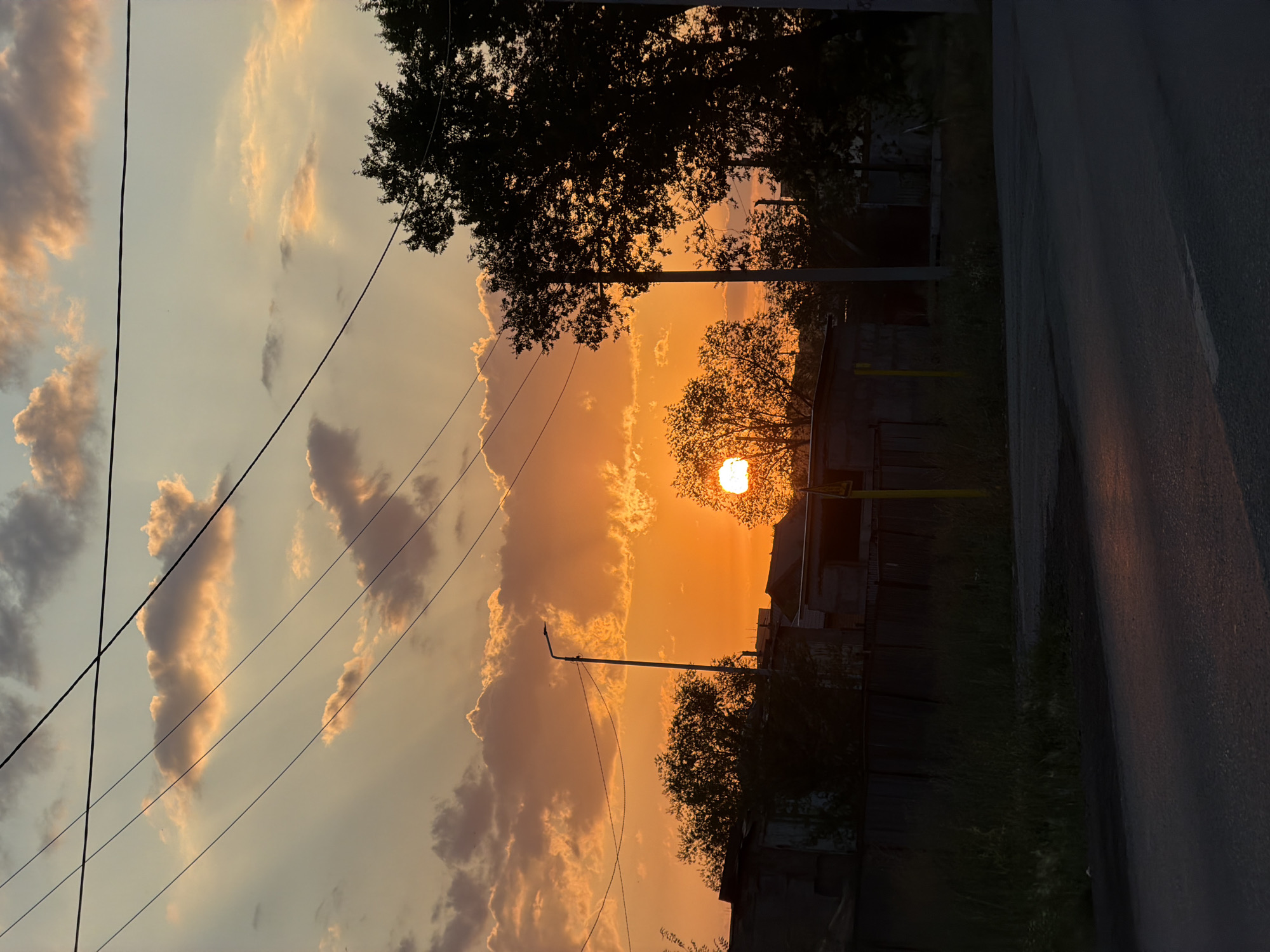 The image captures a sunset in Temirtau, Kazakhstan, with the sky illuminated by a vibrant orange glow. Silhouetted against the dramatic sky are power lines, trees, and a street lamp. In the foreground, a paved road reflects the warm hues of the sunset. Various structures, appearing to be low-rise buildings or sheds, are visible on the left and right sides of the image, casting long shadows. The overall scene depicts a somewhat rural or industrial edge of the city during dusk.