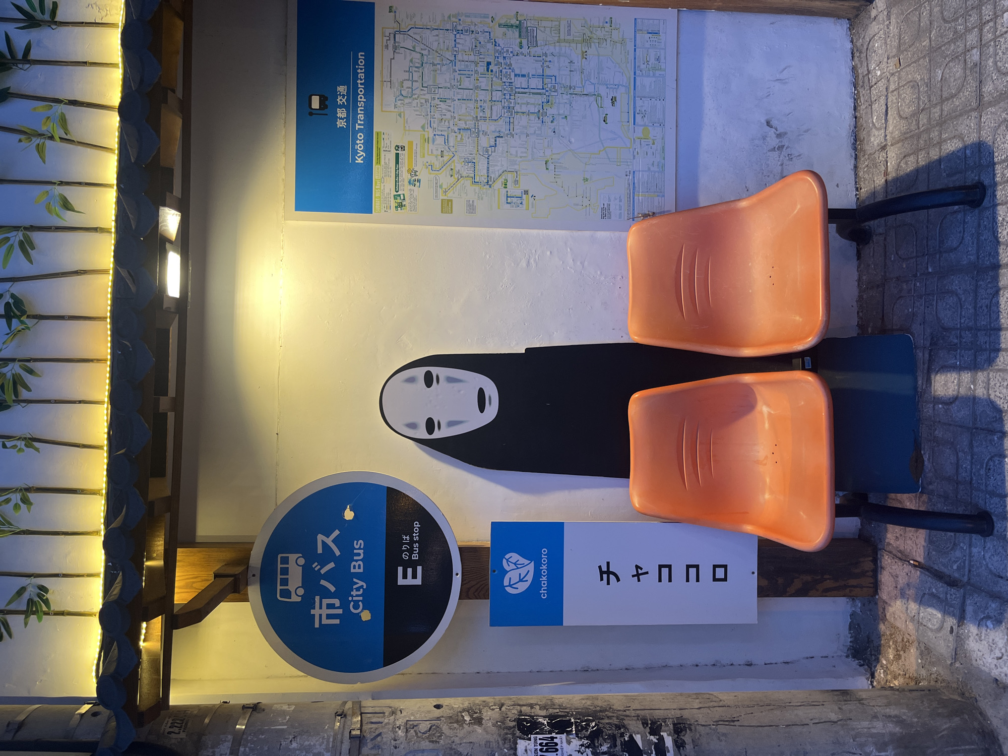 This image depicts a bus stop in the city of Nha Trang, Vietnam, located on a sidewalk next to a building. The bus stop features two bright orange plastic chairs, a sign with the Japanese characters "チャココロ" and the English text "chakokoro" with a leaf logo, and a circular blue and black sign indicating "City Bus" with a bus icon. To the right of the bus stop is a large transportation map of Kyoto, Japan. Behind the chairs and signs, a black silhouette of the character "No-Face" from the animated film Spirited Away is painted on the wall. Above the bus stop signs and the "No-Face" artwork, a decorative element with bamboo stalks and yellow string lights is mounted. The surrounding environment includes a concrete pillar with peeling posters to the left and a tiled sidewalk. The lighting suggests it is either dusk or evening.