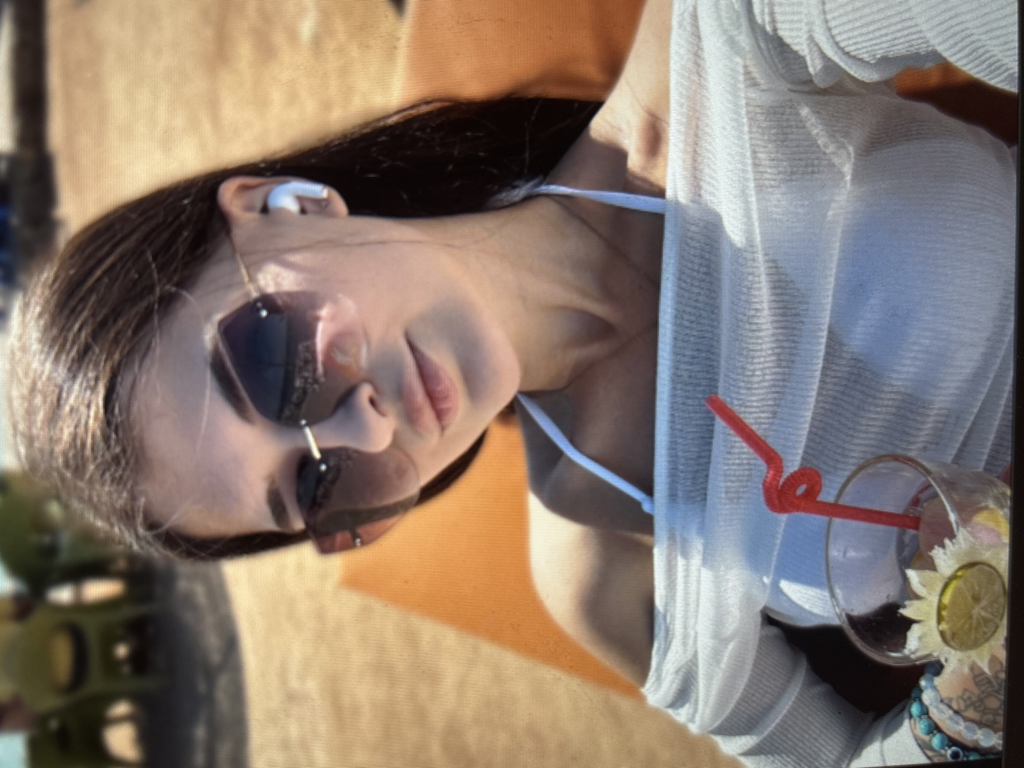 This media file features a young woman relaxing outdoors, likely at a beach or poolside. She is wearing sunglasses and a white, sheer top, with a bikini top visible underneath. A drink with a red straw and a slice of lime sits next to her. She is also wearing wireless earbuds and a bracelet. The overall impression is one of leisure and enjoyment, possibly during a warm, sunny day. The background suggests an outdoor setting with sand or a similar textured surface. The image is likely a casual selfie, given the close-up perspective and the focus on the woman. There is no visible text in the image.