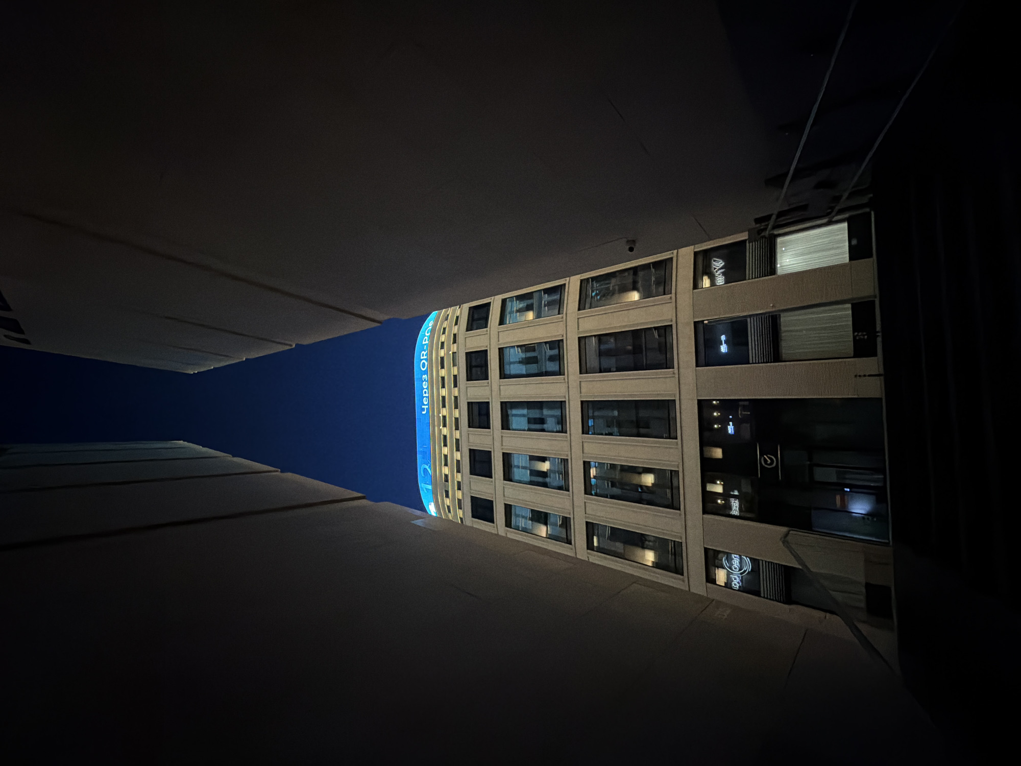 The image captures a nighttime view of a modern, beige-colored office building with numerous rectangular windows. The building is situated in a narrow passage between two dark, imposing structures that frame the scene. The top of the office building is illuminated by a bright blue sign displaying text that appears to be in Cyrillic script, including "Через QR-P". The ground level of the building features entrances and illuminated storefronts, with some windows showing interior lighting. The sky above is a deep, dark blue, indicating it is late evening or night. The perspective is from a low vantage point, looking upwards towards the building. Based on the architectural style and the Cyrillic signage, this location is identified as Minsk, Belarus.