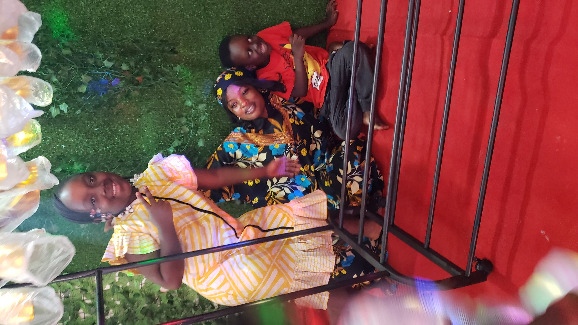In Barden Yana, Nigeria, three individuals are posing for a photograph against a backdrop of artificial greenery and decorative lighting. The scene appears to be set indoors, likely at a celebratory event given the festive decorations.
In the foreground, a young girl, wearing a yellow and white patterned dress with ruffled sleeves and a white beaded necklace, stands and makes a peace sign with her left hand, smiling at the camera.
Behind her, a woman in a dark blue and floral patterned dress with gold trim is leaning back and smiling, her head tilted. Next to her, to the right, a young boy in a red t-shirt and dark pants is seated, looking towards the camera with a wide, slightly strained smile.
The individuals are positioned on a red carpeted surface in front of a dark metal structure, possibly a rack or a display unit with multiple horizontal bars. The lighting in the image is mixed, with a soft glow from the decorations and some noticeable lens flare. The overall impression is of a candid moment captured during a gathering.