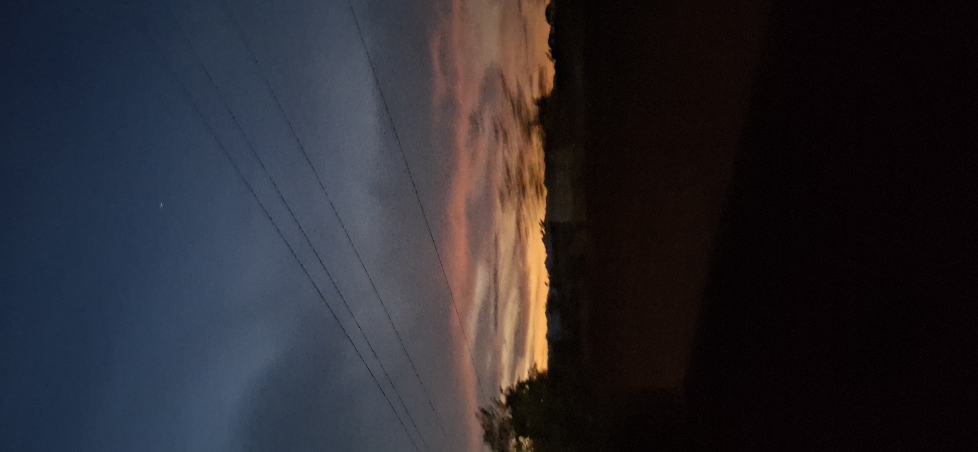 The image shows a sunset sky with a dark foreground. In the upper left, the sky is a deep blue, transitioning to a band of pink, orange, and white clouds towards the center. Silhouetted power lines run across the left and center of the frame. On the right side, a large dark area obscures the view, possibly a building or dense foliage. The lower part of the image is obscured by darkness, but some foliage is barely visible. The image is shot vertically. Without specific information about the environment, it is impossible to accurately determine the location in Vallecito, Venezuela.