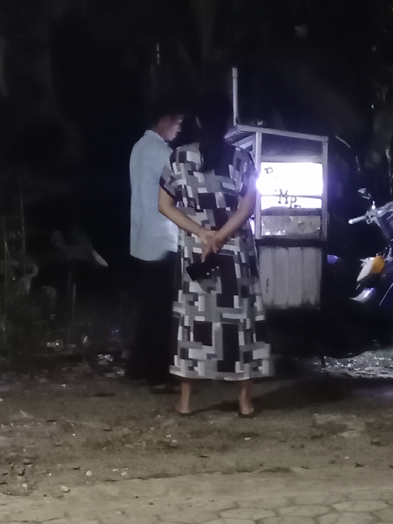 In Parage, Indonesia, at night, two individuals stand outdoors. A person on the left wears a light blue long-sleeved shirt and dark pants. A person on the right wears a dress with a geometric pattern of black, white, and gray squares and rectangles. This person has their hands clasped behind their back. Behind them is a lighted kiosk with a motorcycle parked to the right. The ground is composed of dirt and small stones.