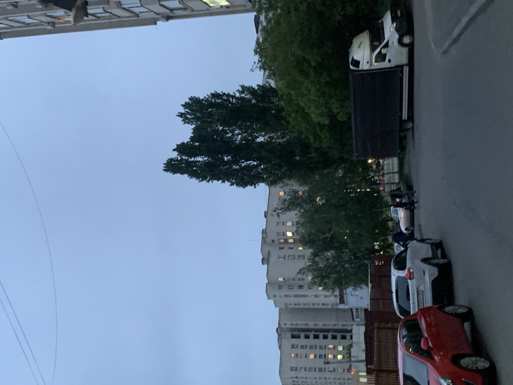 The image depicts a street scene in Stavropol, Russia, at dusk. Several buildings with lit windows are visible, suggesting residential areas. A variety of vehicles are parked along the side of the street, including a red car, a white car, and a large box truck. Trees are present alongside the street. There are several people are standing around parked cars. The lighting suggests it is twilight or shortly after sunset. The overall impression is of a quiet urban environment in the evening.