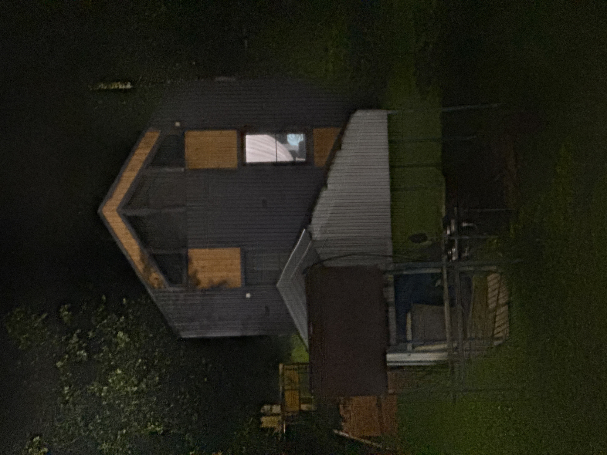 In the city of Doma 1096 Km, Russia, at night, a modern, dark-paneled house stands partially obscured by trees. The house features a peaked roof with lighter wood trim and several rectangular wood accents. One window on the upper floor emits a soft white light, revealing a white curtain and a blurred figure inside. In the foreground, a lower structure with a slanted, corrugated metal roof extends from the main house, supported by metal poles. This structure appears to be a covered patio or extension. Beneath this, a dark rectangular object, possibly a bench or table, is visible, along with a blue container and other assorted items. The ground is covered in grass, and the overall scene is dark, suggesting it was taken in low light conditions.