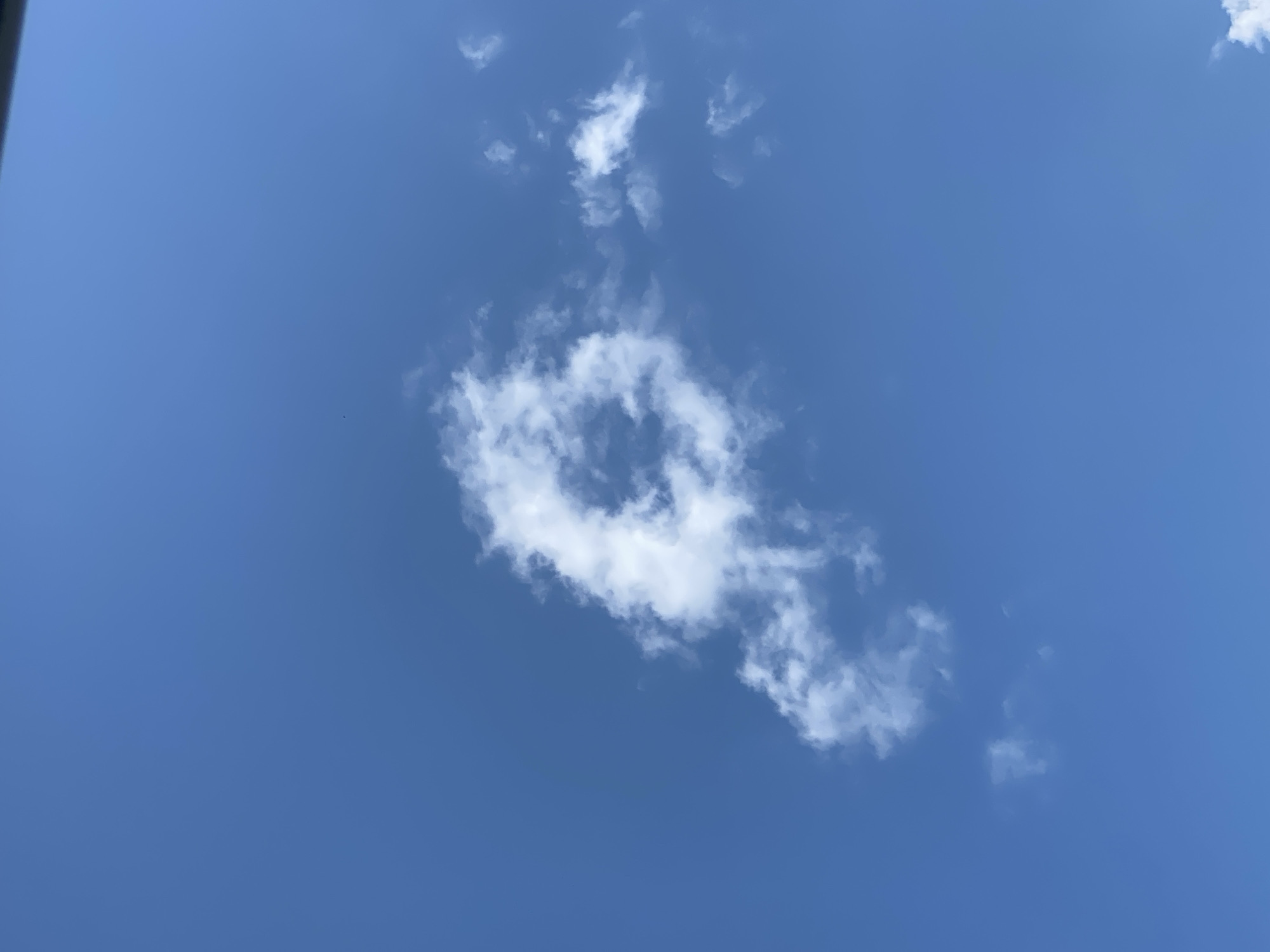The image shows a blue sky with scattered white clouds. One cloud, located near the center of the frame, has a distinct ring-like shape with wispy extensions. The location is not specified.