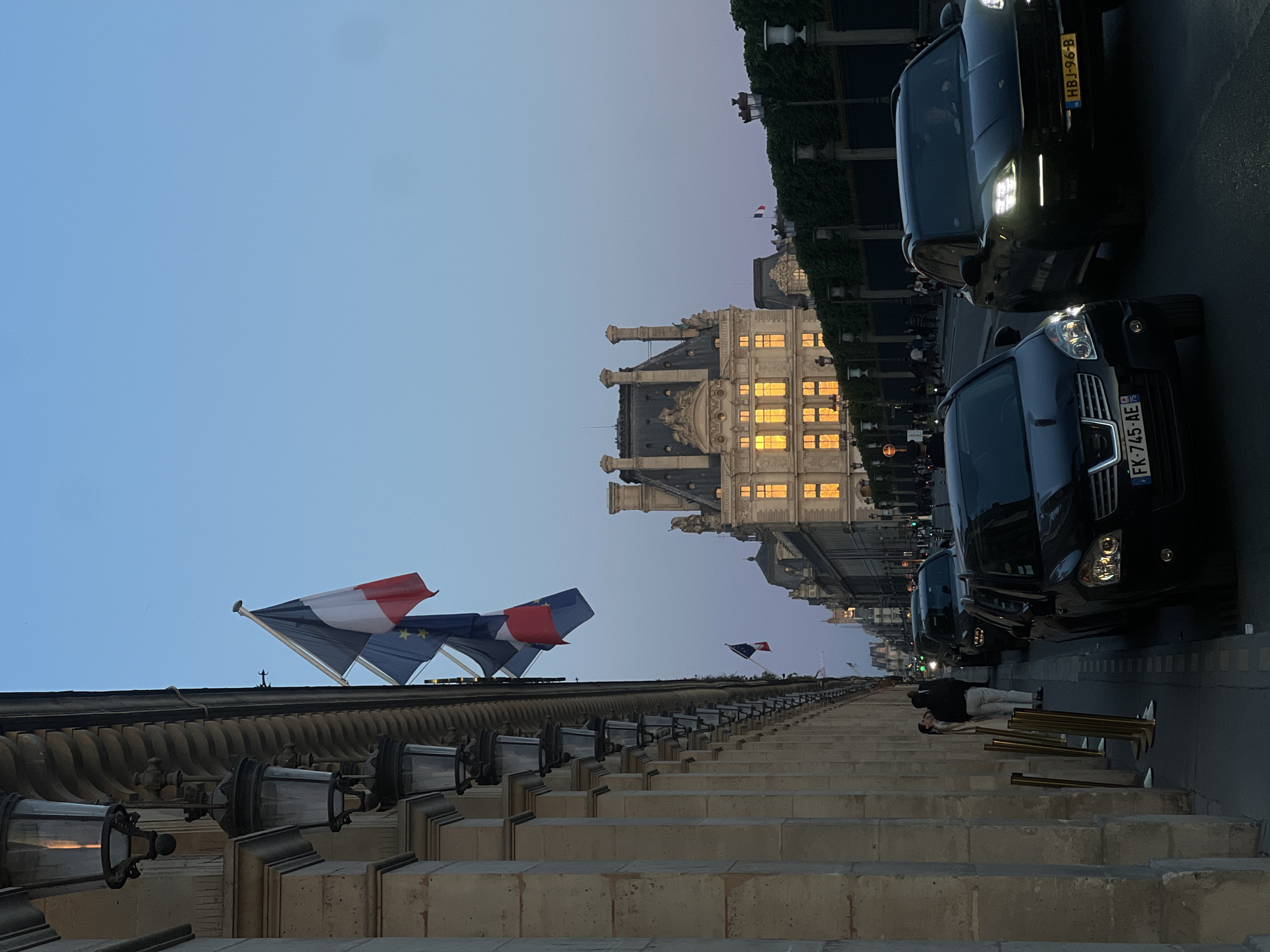 The image depicts an urban street scene at dusk or early night, characterized by grand architecture and a tranquil atmosphere.
In the foreground, running along the left side, is a long, stone structure resembling a series of steps or a grand balustrade, adorned with ornate, dark lampposts. From this structure, prominently fly a French flag (blue, white, red vertical stripes) and a European Union flag (blue with a circle of yellow stars). Further along the structure, another smaller French flag is visible. Near the base of these steps, a person dressed in light pants and a dark jacket is lying on their side on the pavement, next to several golden stanchions.
To the right of the stone steps, a street is visible with several dark-colored cars parked. The car closest to the viewer displays a French license plate "FK-745-AE". Behind it, another car features a Dutch license plate "HBJ-96-B". The cars' headlights are on, indicating low light conditions.
In the background, on the right side of the image, stands a magnificent, multi-storied building with intricate architectural details, including ornate carvings and chimneys. Many of its windows are brightly illuminated with a warm, golden light, highlighting its grand facade. Other blurry figures can be discerned walking on the street near this building.
The sky above is a clear, gradient blue, transitioning from a lighter hue at the horizon to a deeper indigo overhead, typical of twilight. The combination of the French and EU flags, the French license plate, and the distinctive European architecture strongly suggests the location is a major European capital, likely Paris. No specific event is taking place; it's a serene street view.