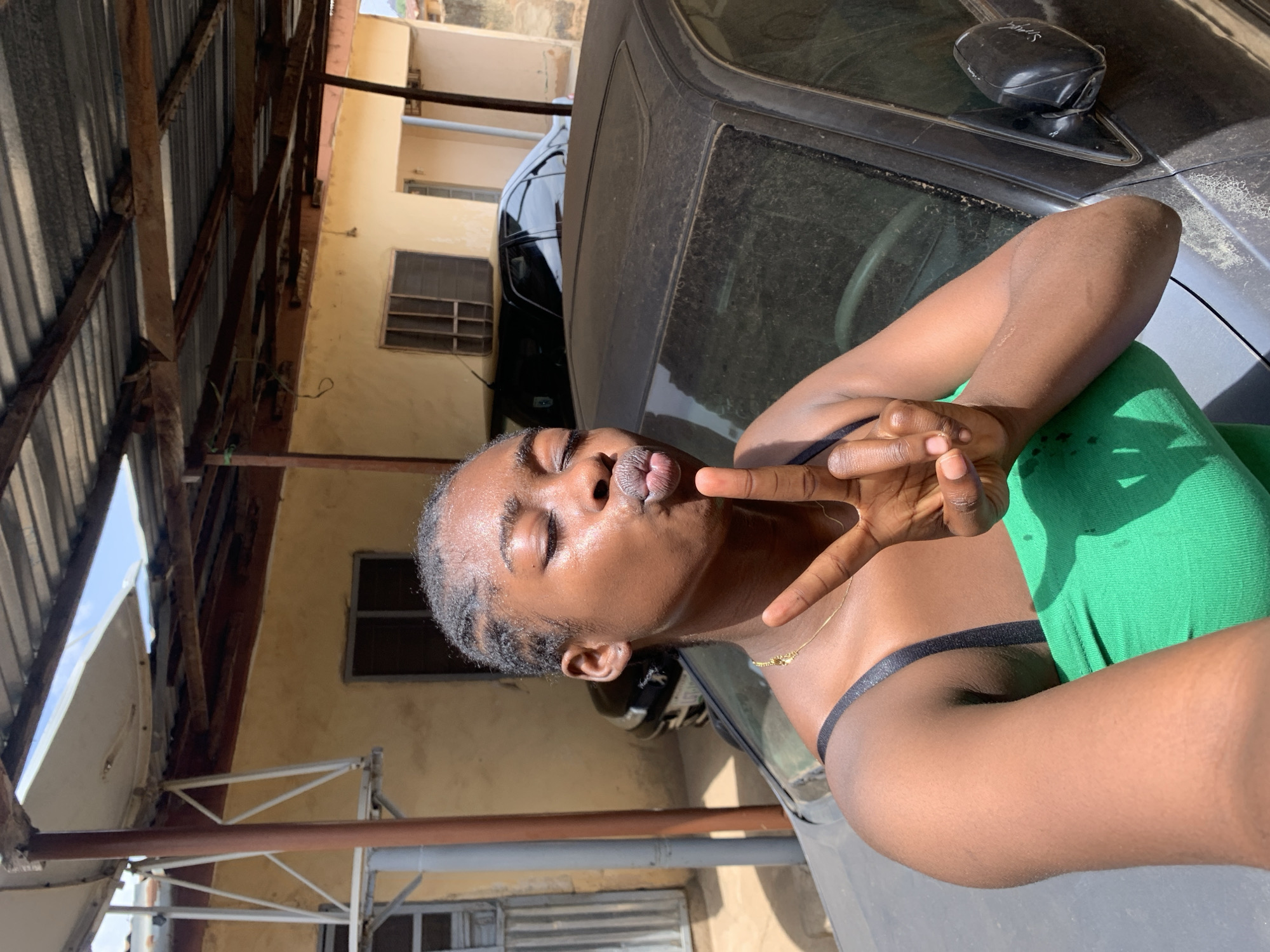 A young, dark-skinned woman is taking a selfie outside in Rafin Par, Nigeria. She is pouting her lips and holding up a peace sign. She is wearing a green top with black straps. She appears to be sitting on or leaning against a dusty black car. The background includes a tin roofed structure, beige walls, and windows.